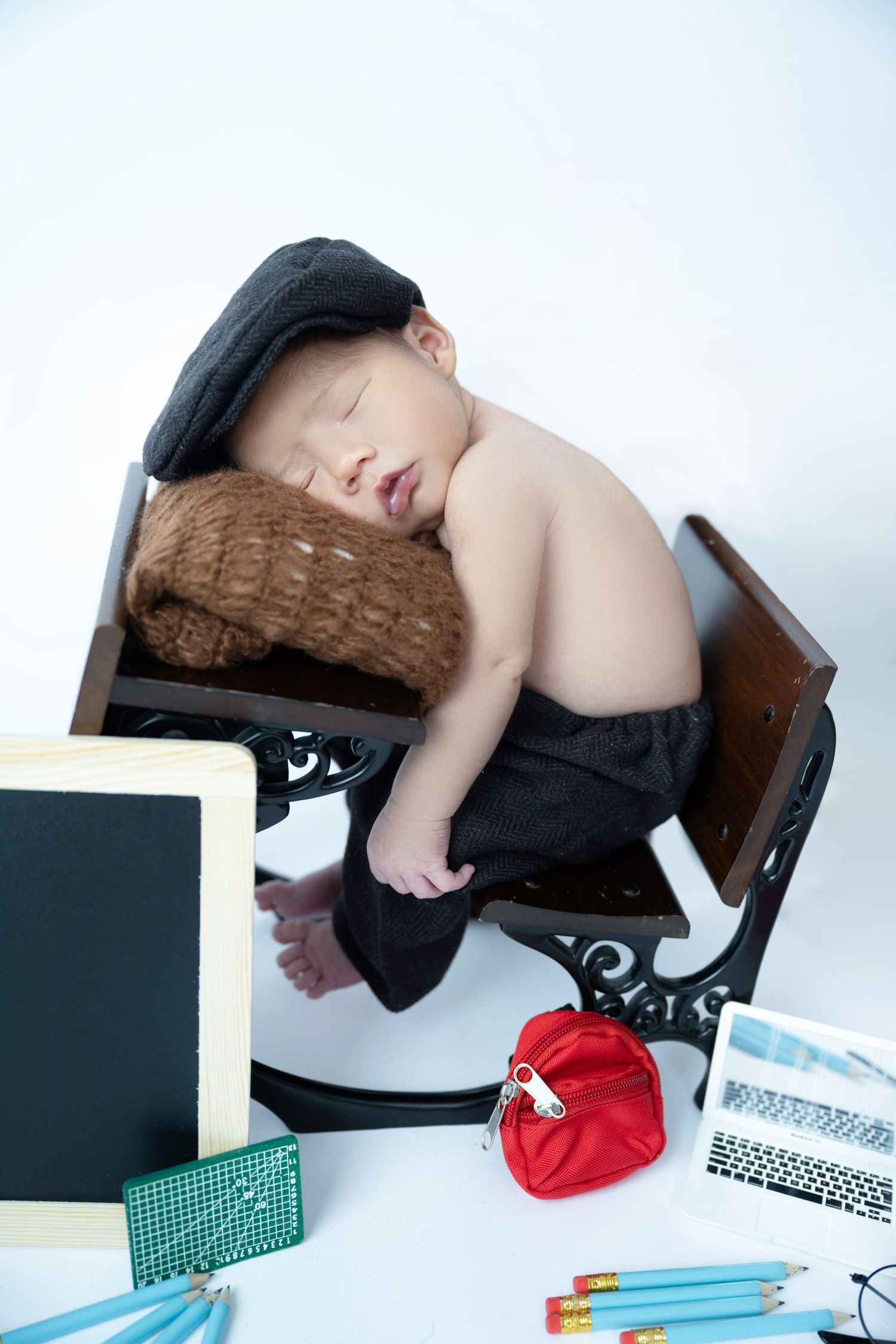 Sleeping newborn baby wearing a newsboy cap posed on a vintage school desk with pencils and a chalkboard.
