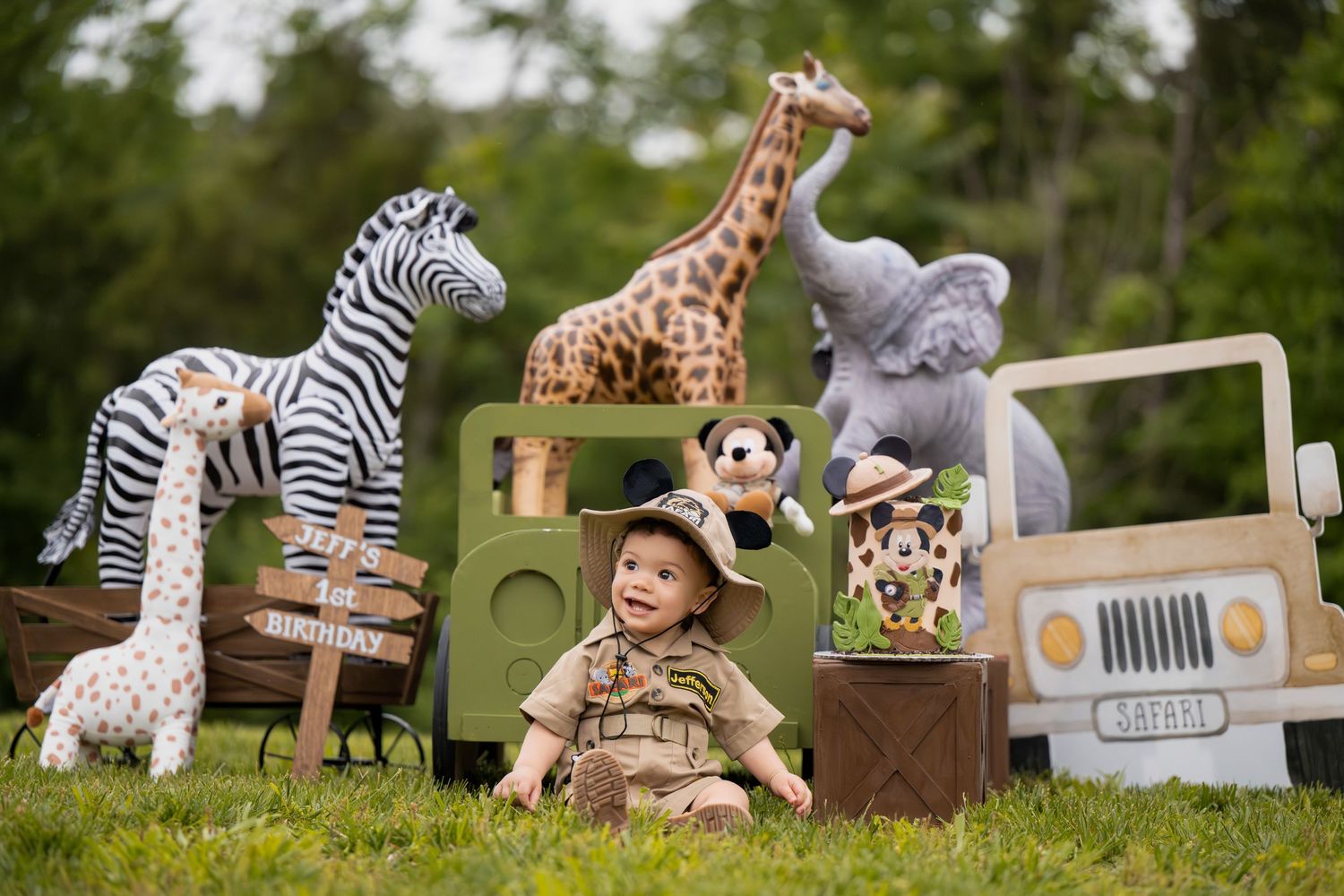 Baby in safari outfit sits on grass surrounded by stuffed animal toys and a toy jeep outdoors.