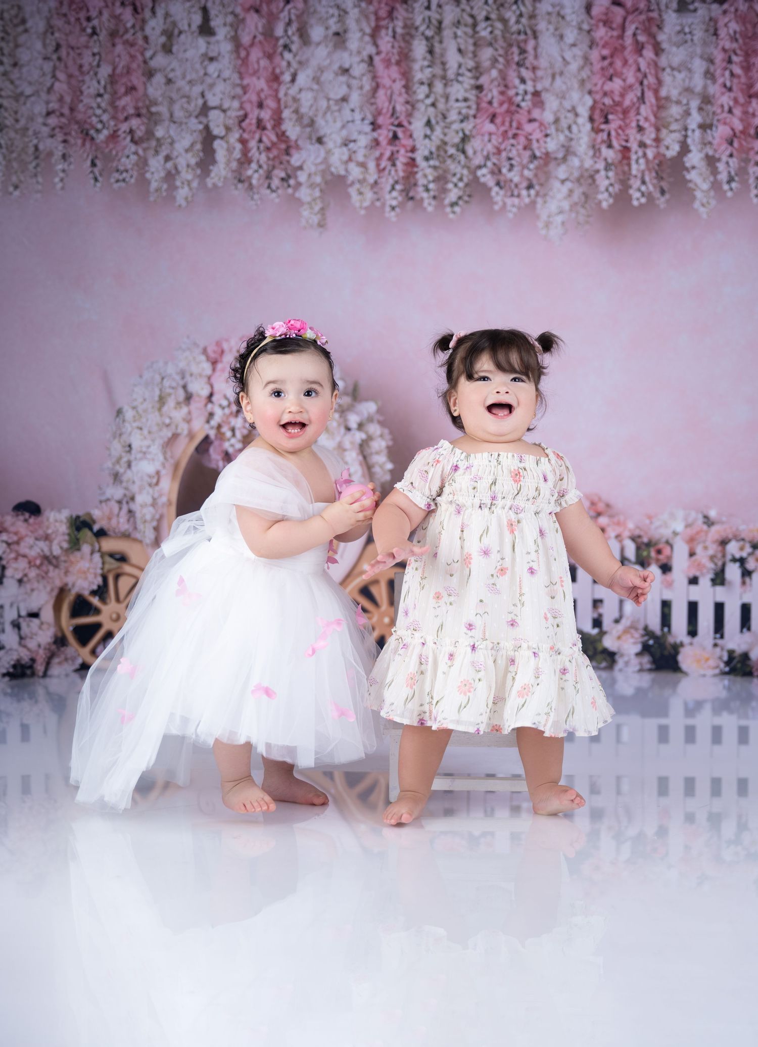 Two toddlers in white dresses play against pink floral backdrop with white picket fence.