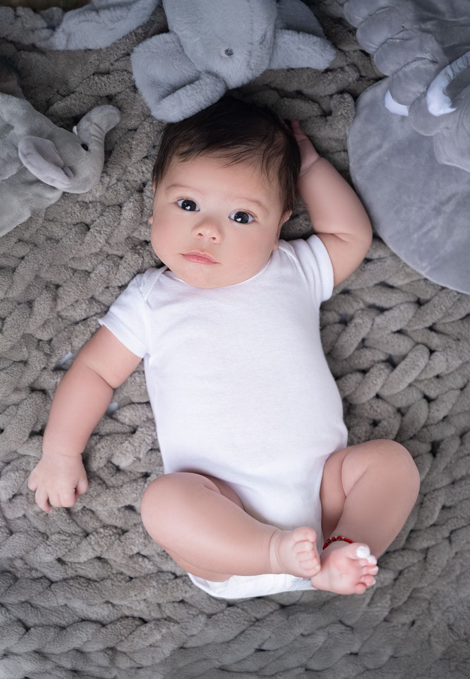 Baby in white onesie lying on grey knit blanket with plush toys.