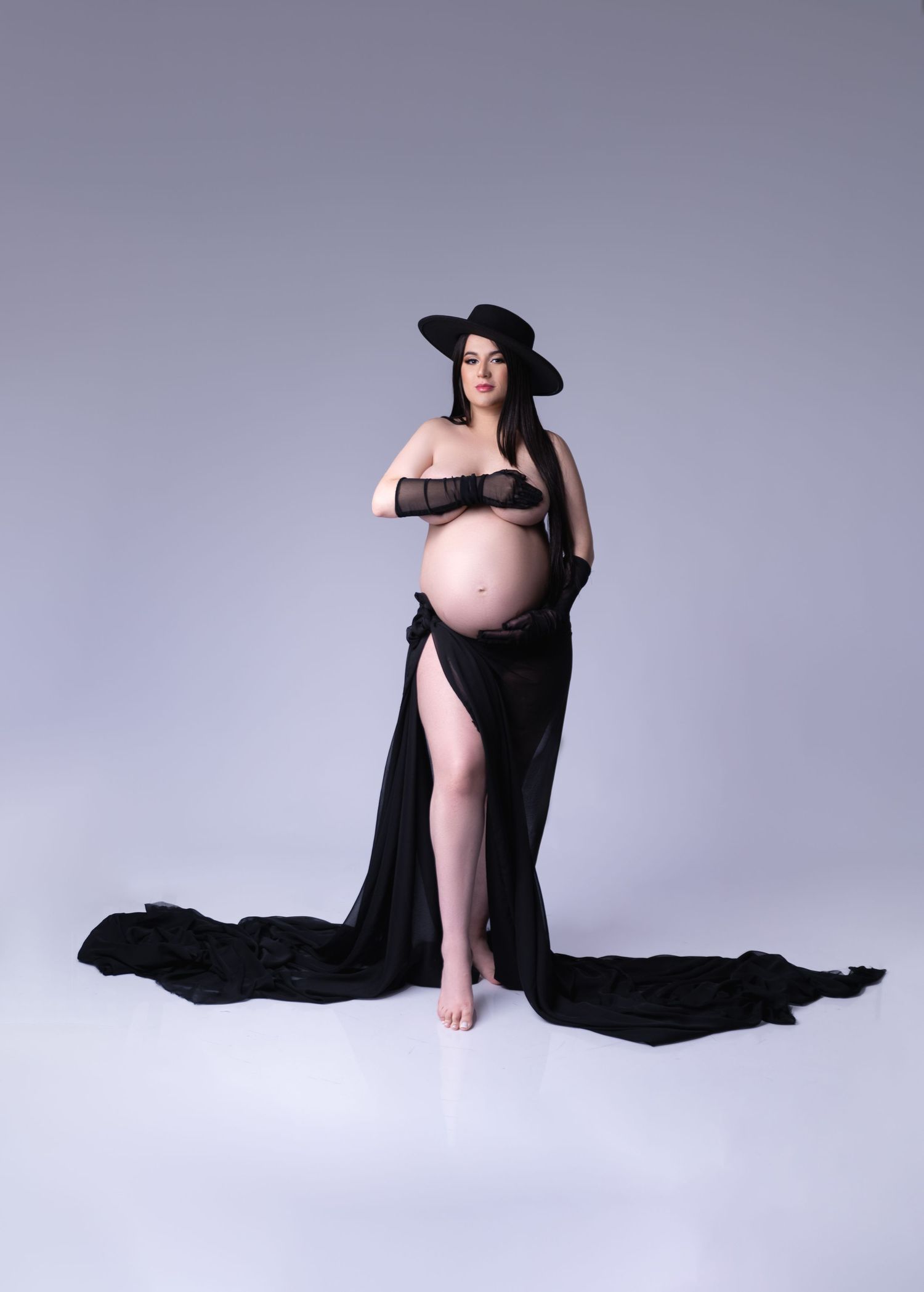 Artistic maternity portrait in black flowing fabric and wide-brimmed hat against neutral background.