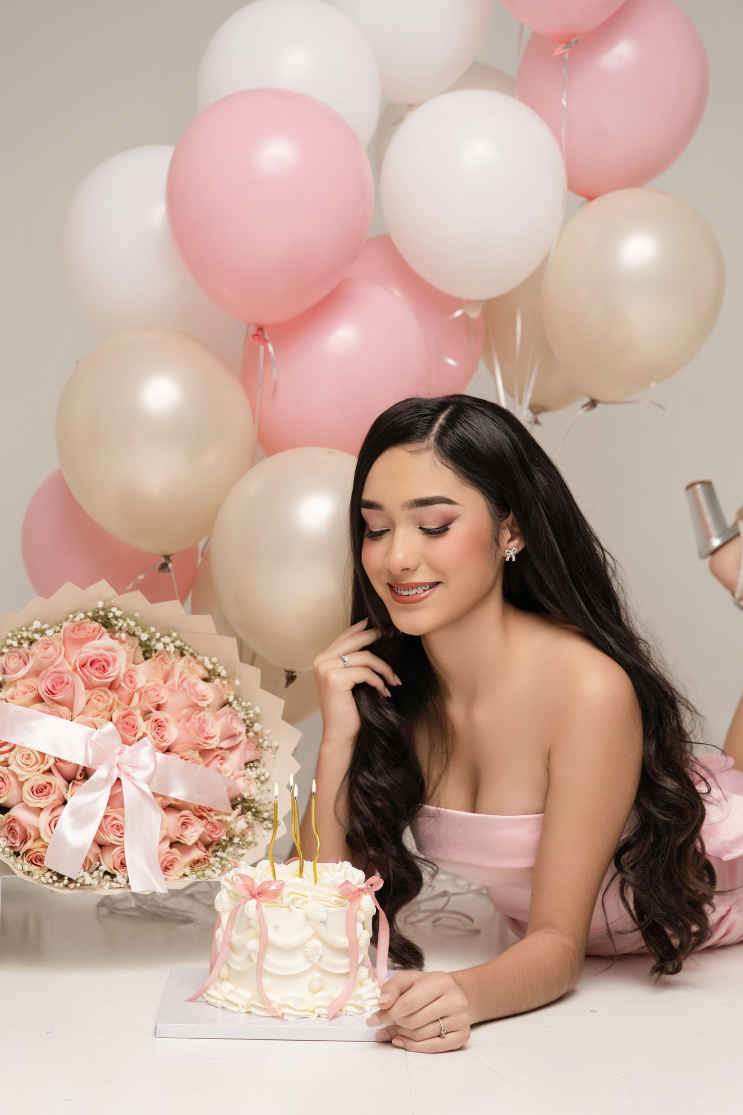 Young woman in pink dress smiles at birthday cake surrounded by pink and white balloons and a rose bouquet.