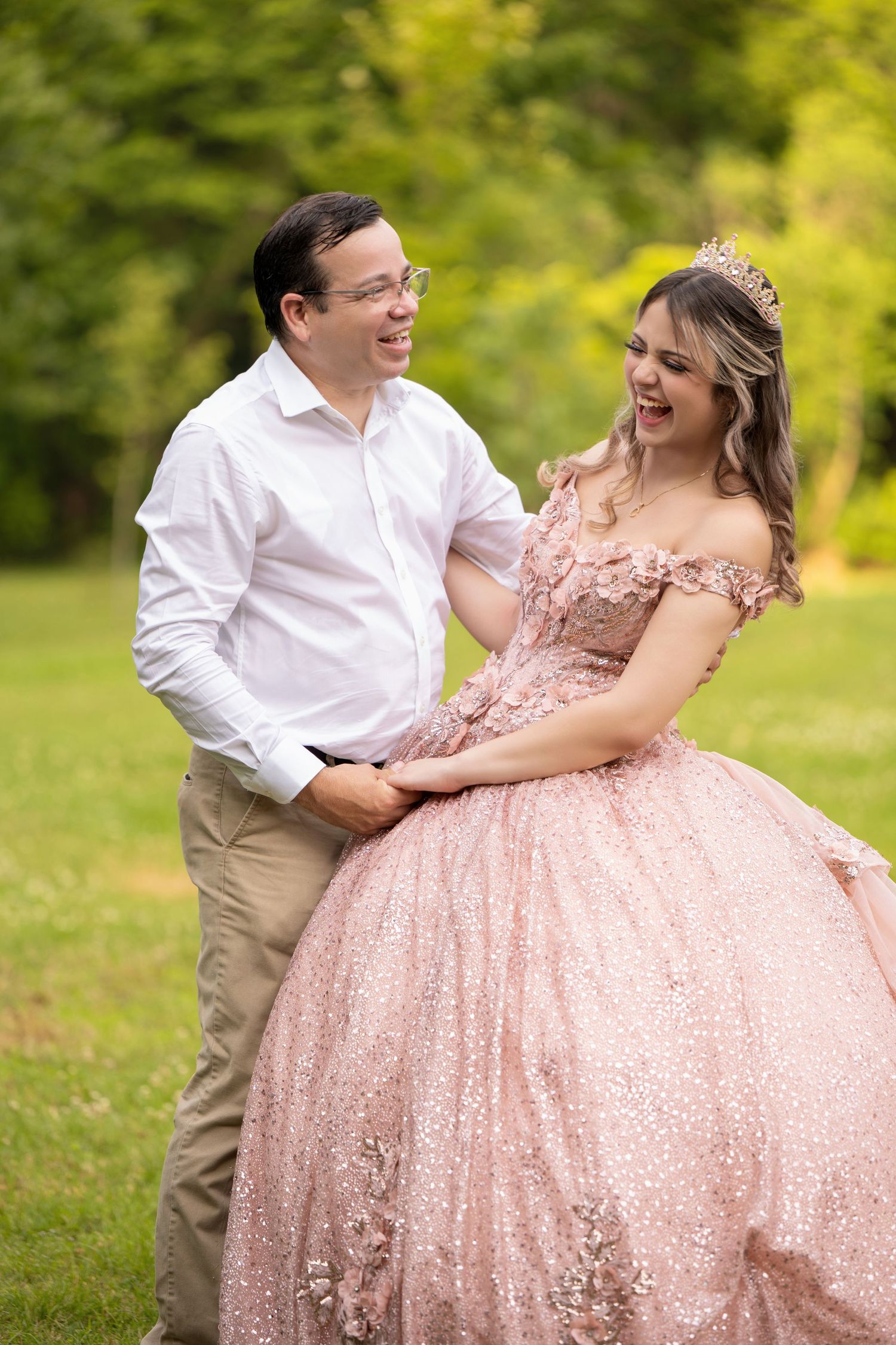 A joyful couple laughs together outdoors, the young woman wearing a sparkling pink ball gown and tiara for her quinceañera.