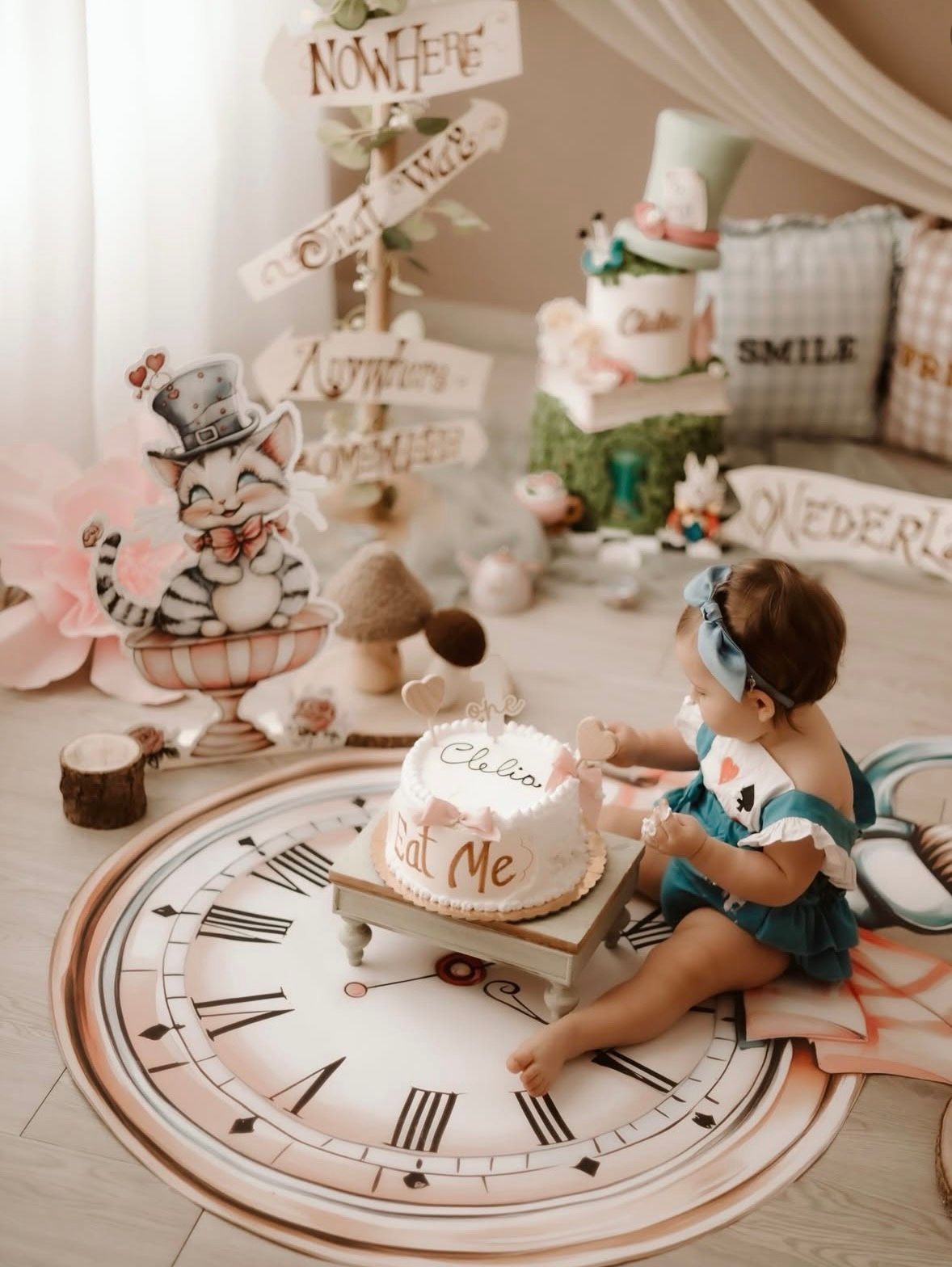 Baby girl sits on clock rug with Alice in Wonderland themed first birthday cake smash setup and whimsical decorations.