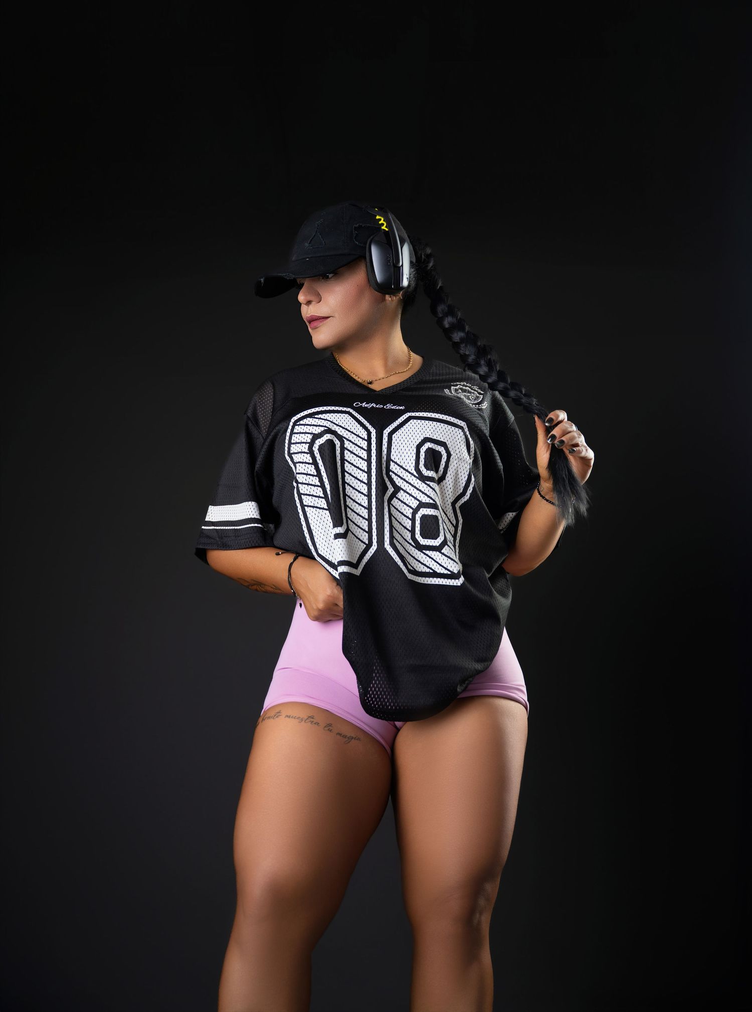 Woman in a black cap, headphones, number 08 football jersey and pink shorts posing with a long braid against a dark studio backdrop.