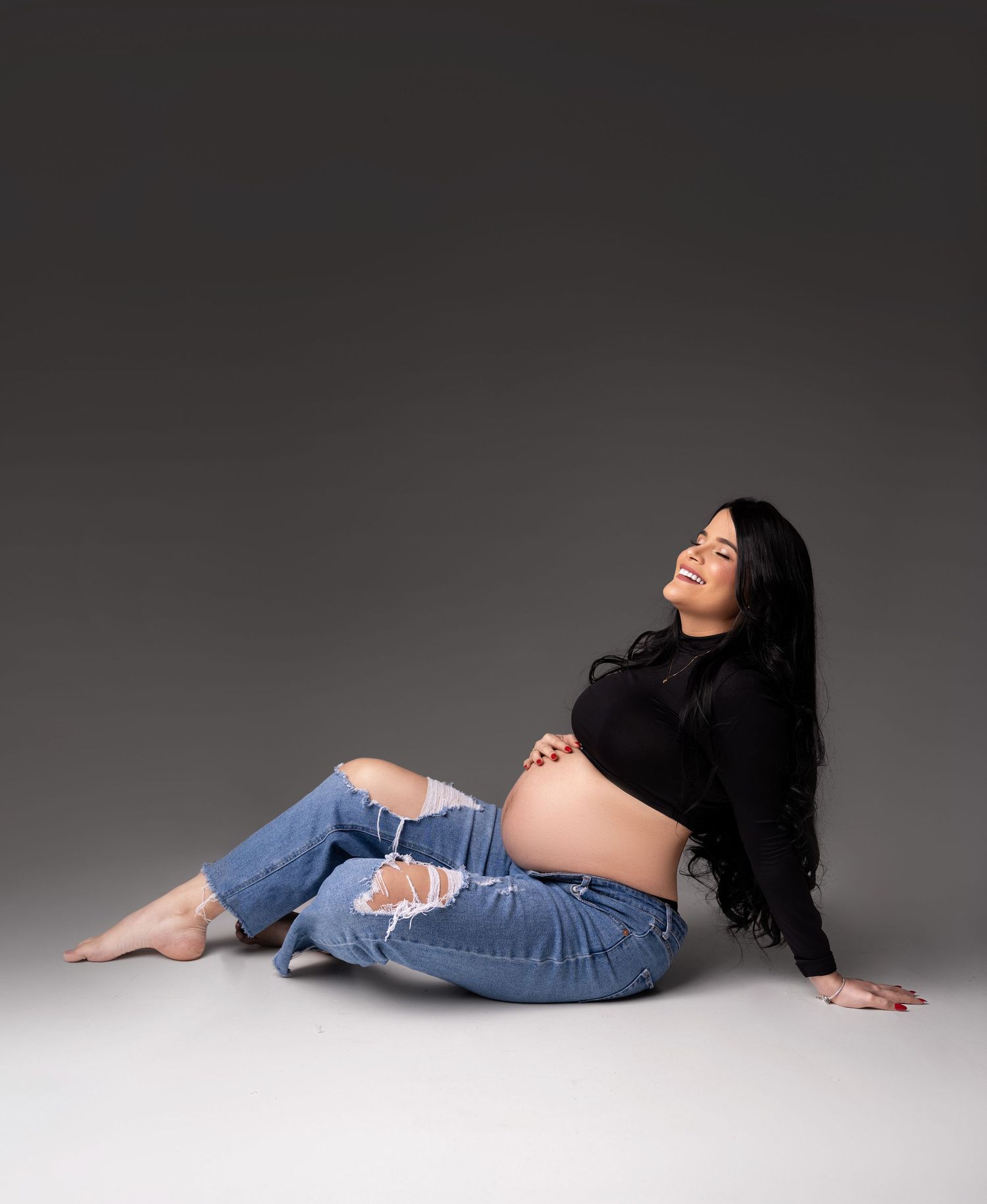 A pregnant woman in ripped jeans and black sweater reclines on the floor during a maternity photoshoot with gray backdrop.
