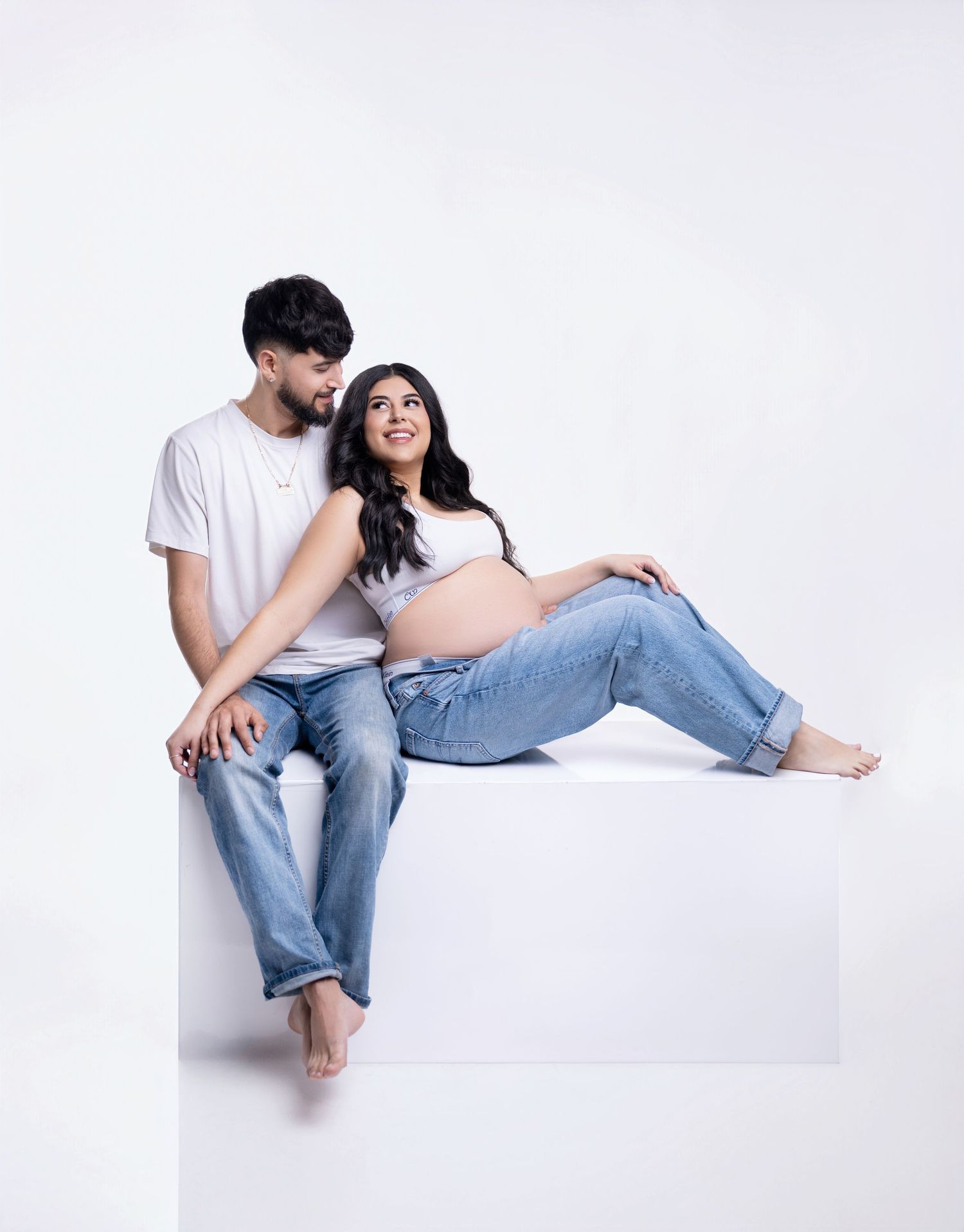 A couple in casual attire sits together on a white background in a series of intimate, playful poses.