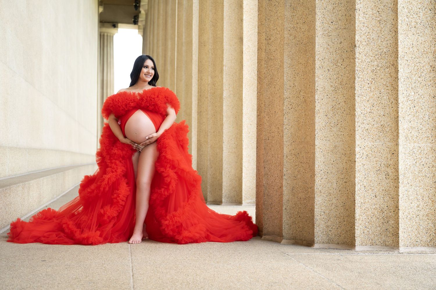 A pregnant woman poses in a flowing red gown against stone columns in an elegant maternity photoshoot.