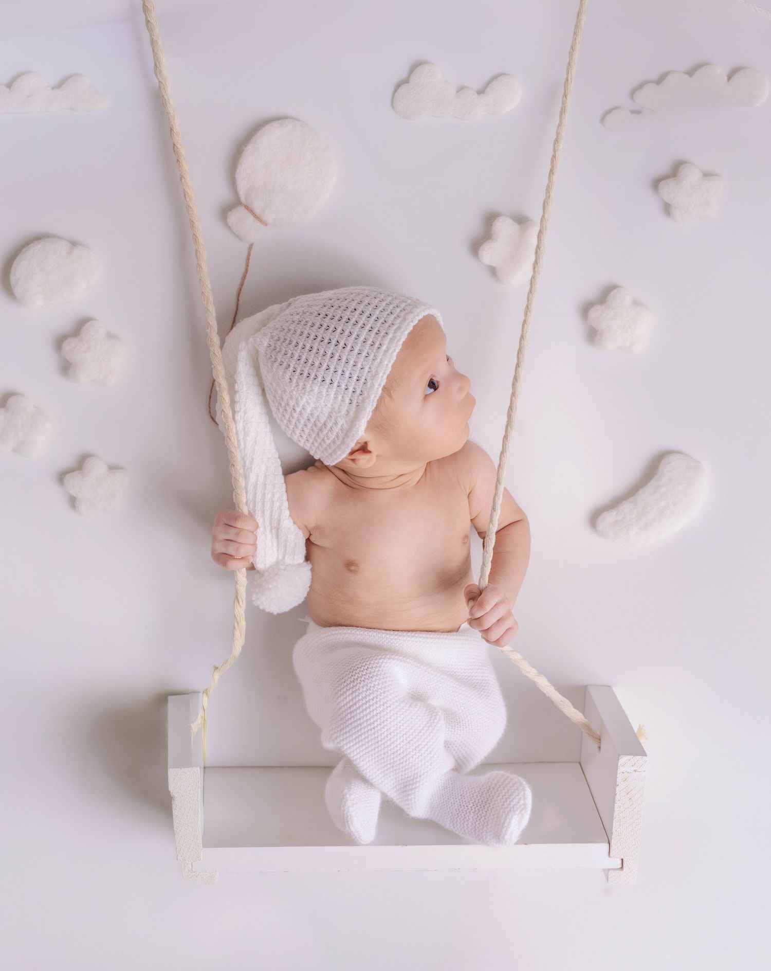 Adorable newborn baby in white knit hat sitting on a swing surrounded by cloud decorations.