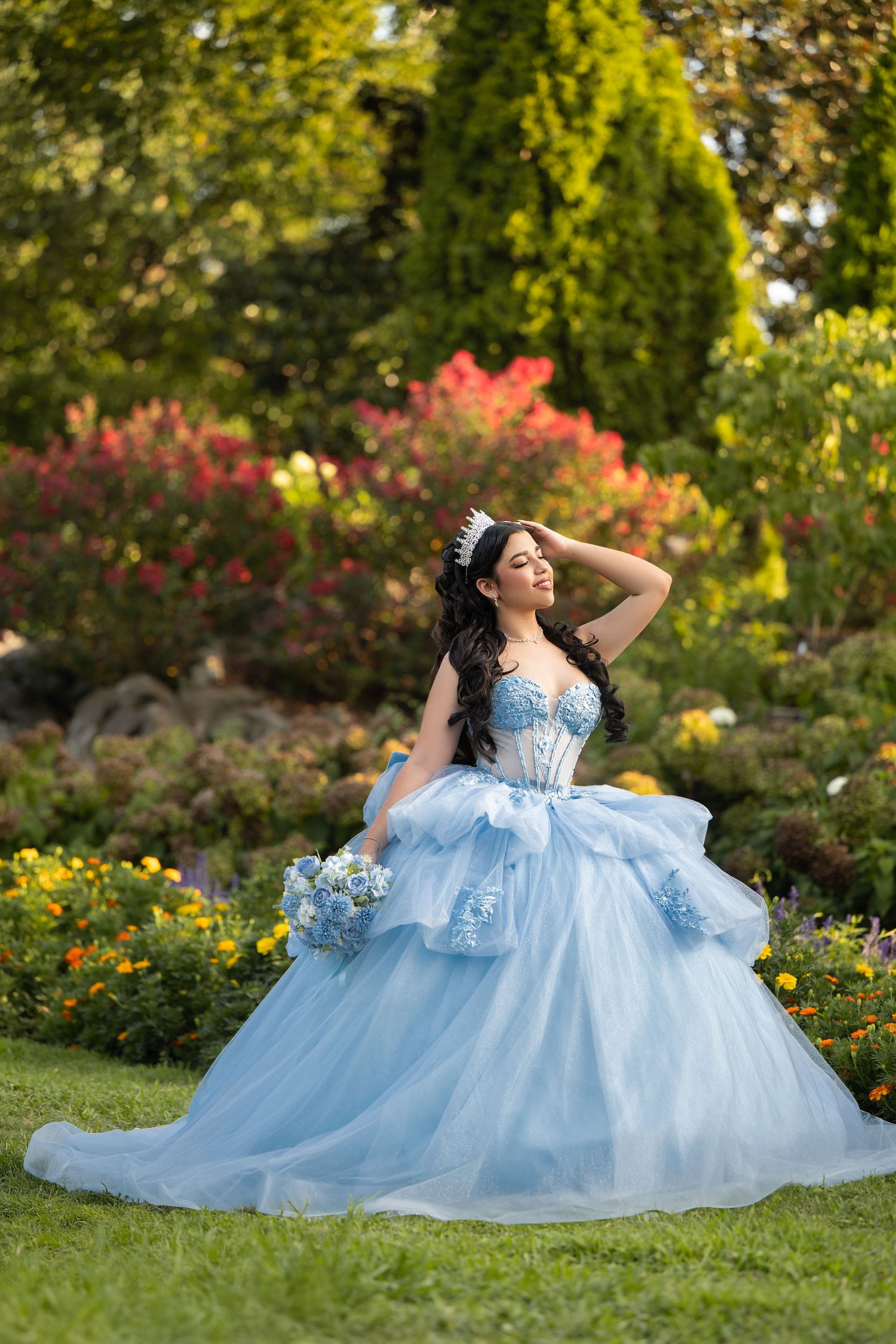 Young woman in a light blue ball gown and tiara poses gracefully in a colorful garden holding a bouquet.