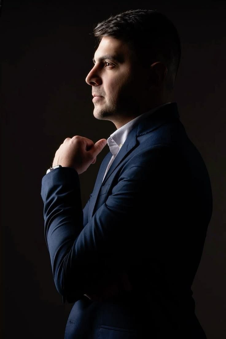 A professional man in a navy blue suit poses thoughtfully against a dark background in a dramatic side profile portrait.
