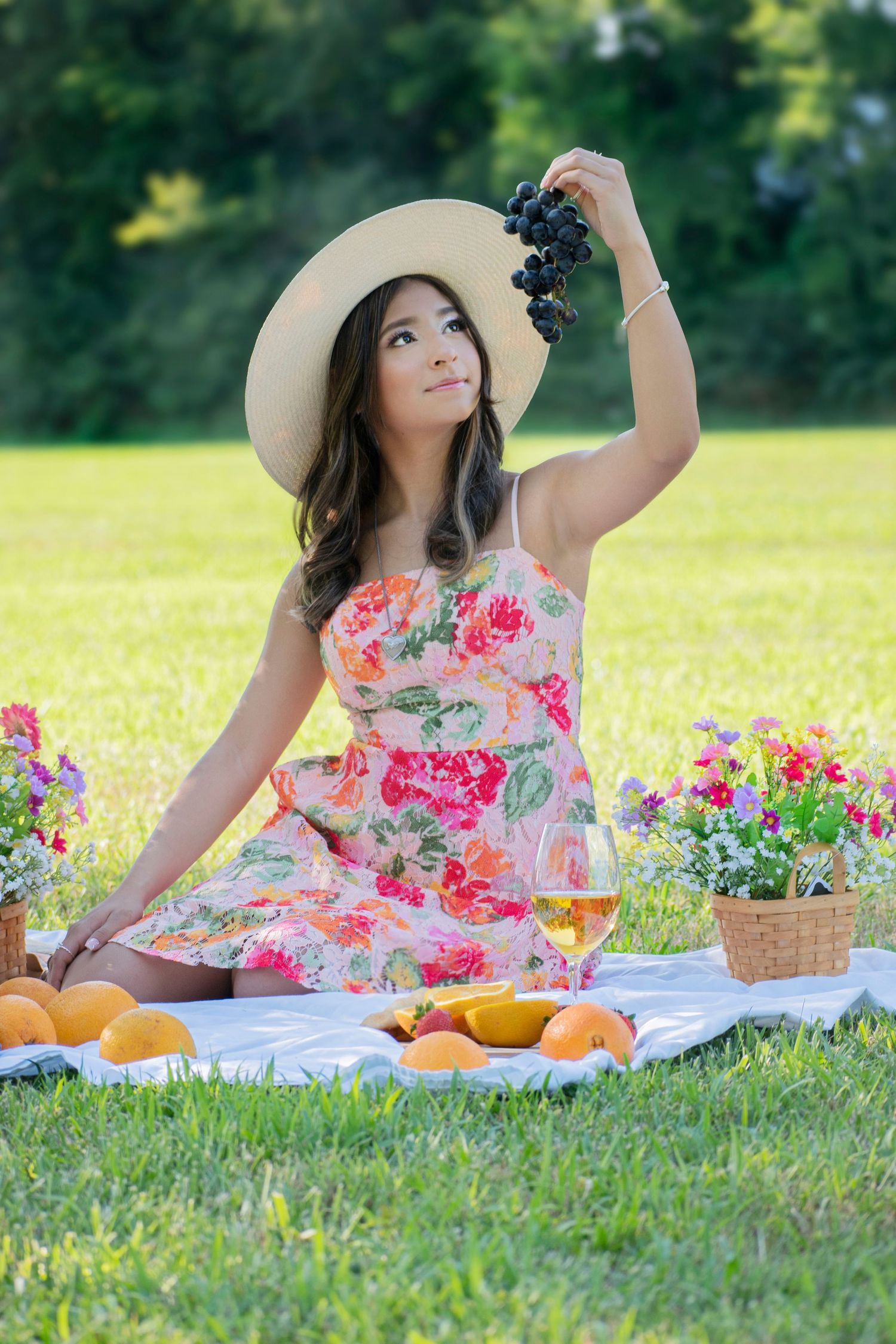 Young Asian woman in floral dress and sun hat holds grapes at a sunny outdoor picnic with fruit, wine, and flowers.