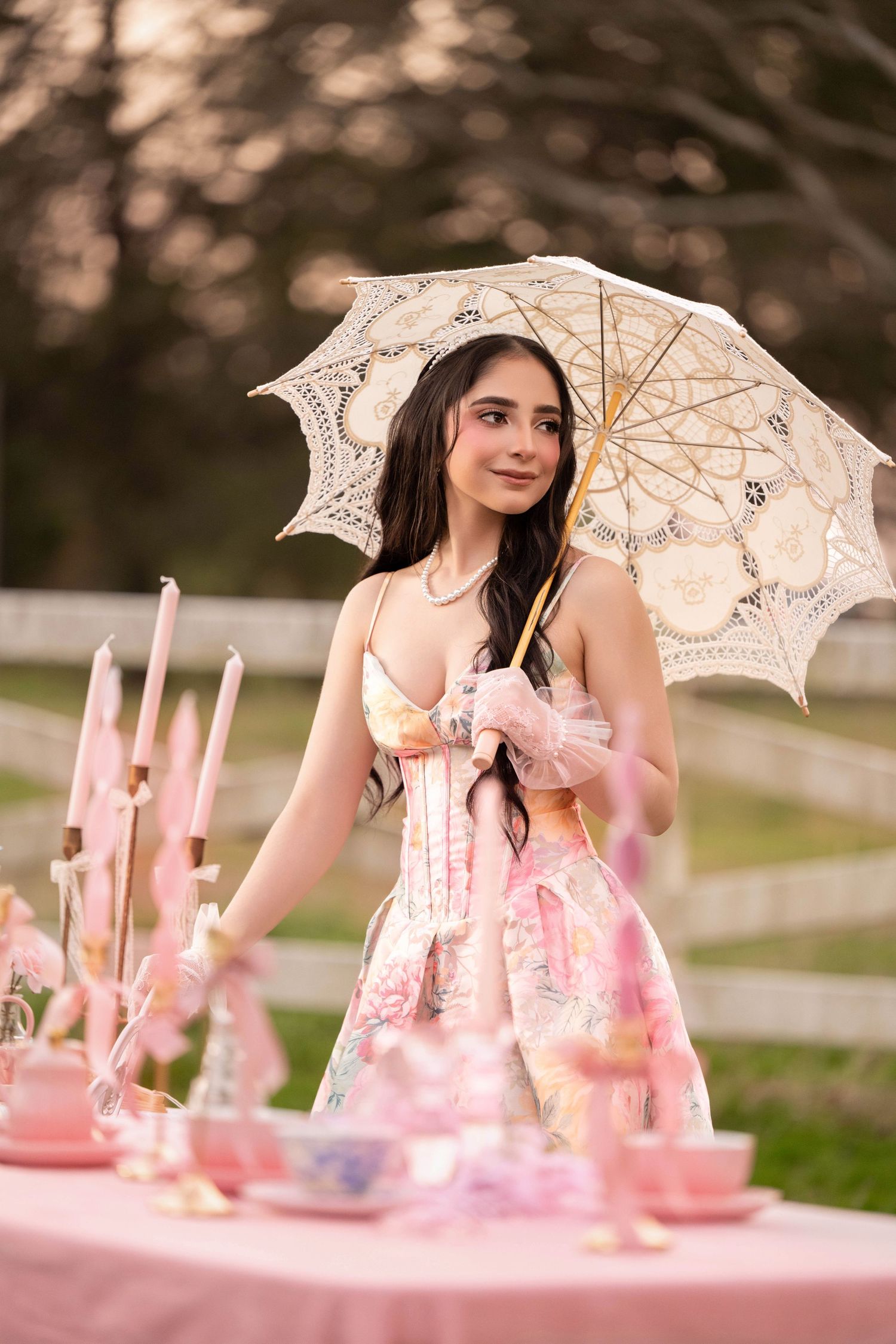 Young woman in pink floral dress holding lace parasol at elegant outdoor pink tea party table setting.