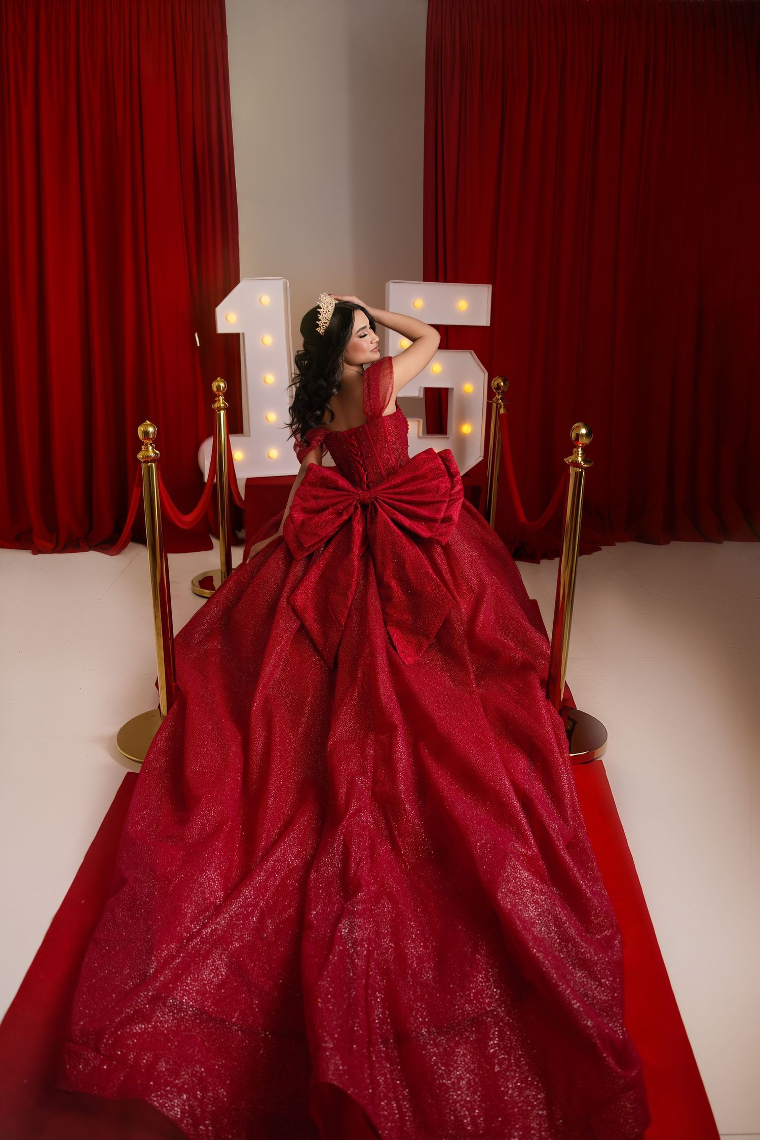 Girl in red ballgown with large bow poses on red carpet in front of illuminated number 16 sign.