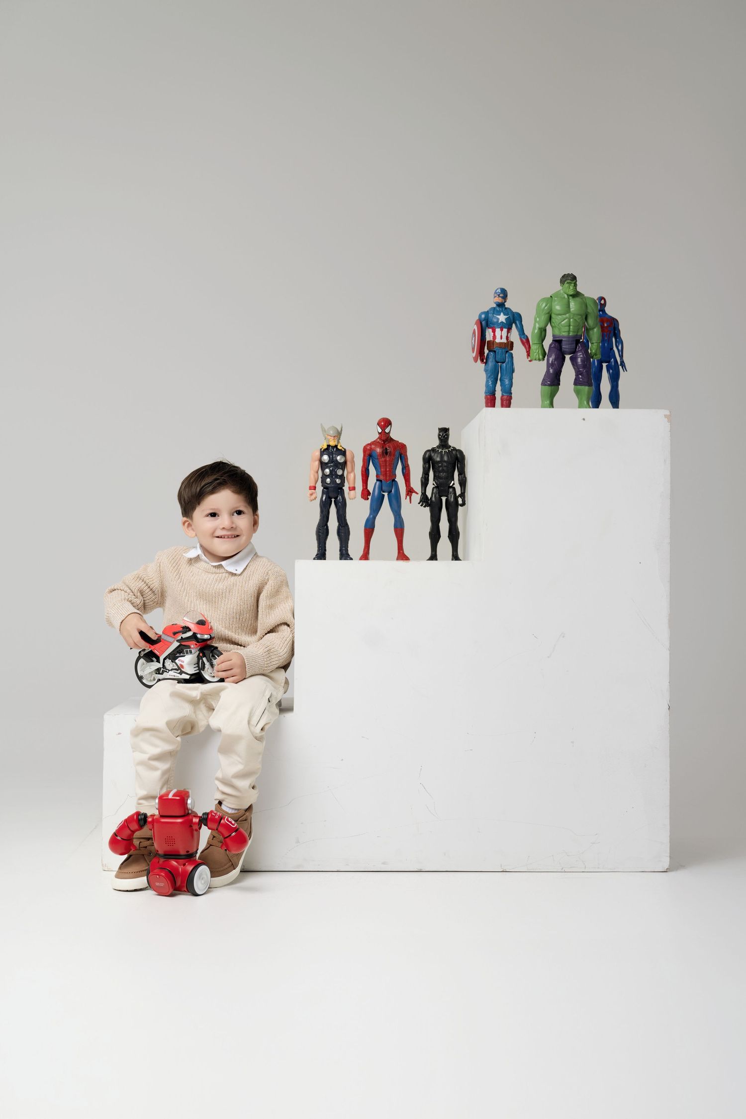 Smiling boy holding toy motorcycle with Marvel superhero action figures on white steps.