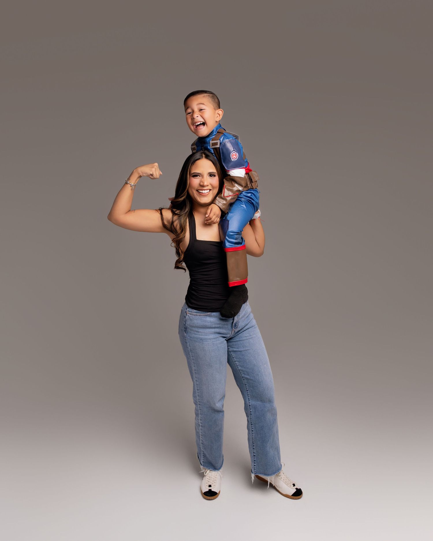 A woman in a black tank top and jeans flexes her arm while a young boy in a superhero costume sits on her shoulders.