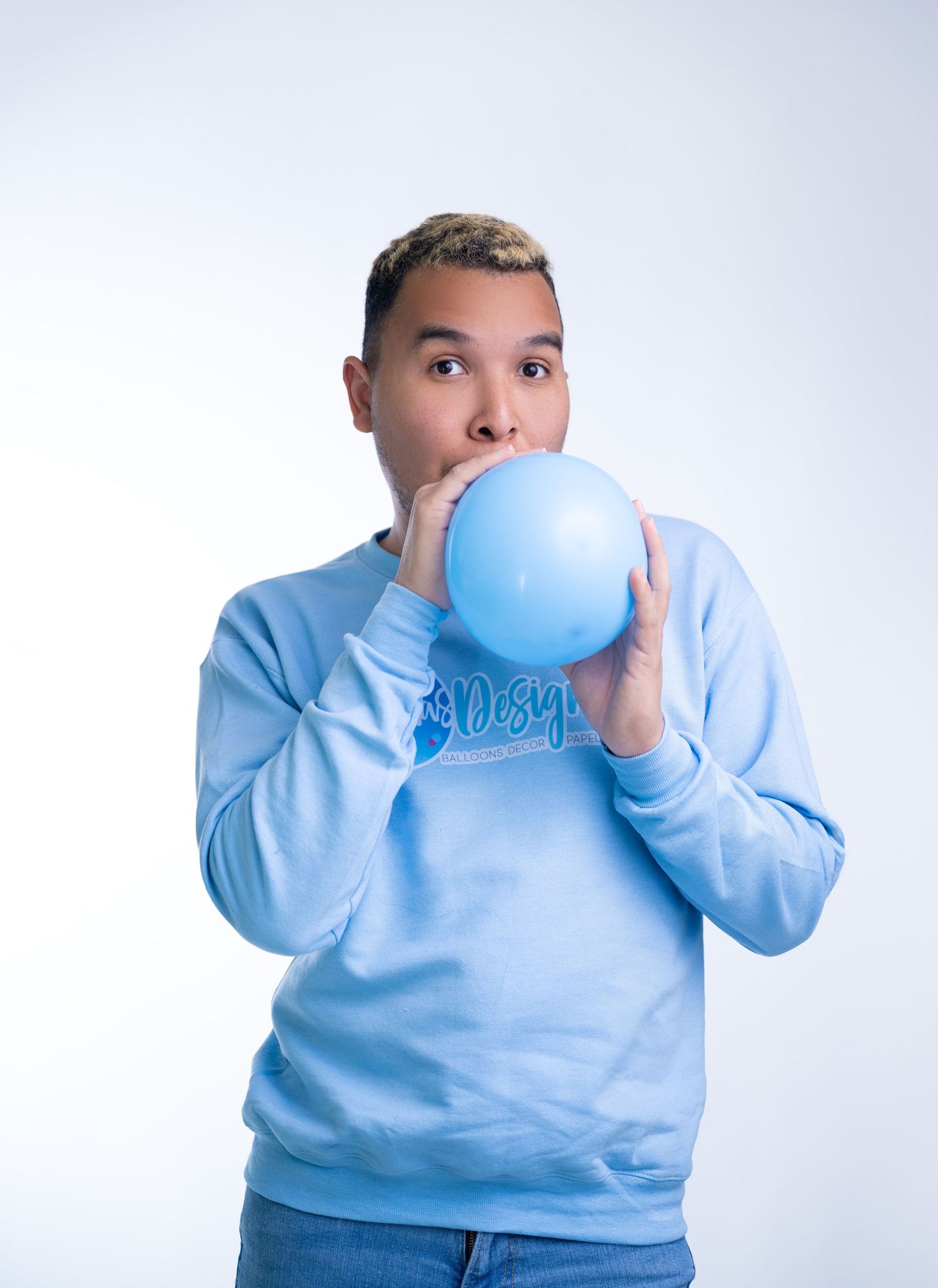 Young man in light blue sweatshirt blowing up a blue balloon against a white background.