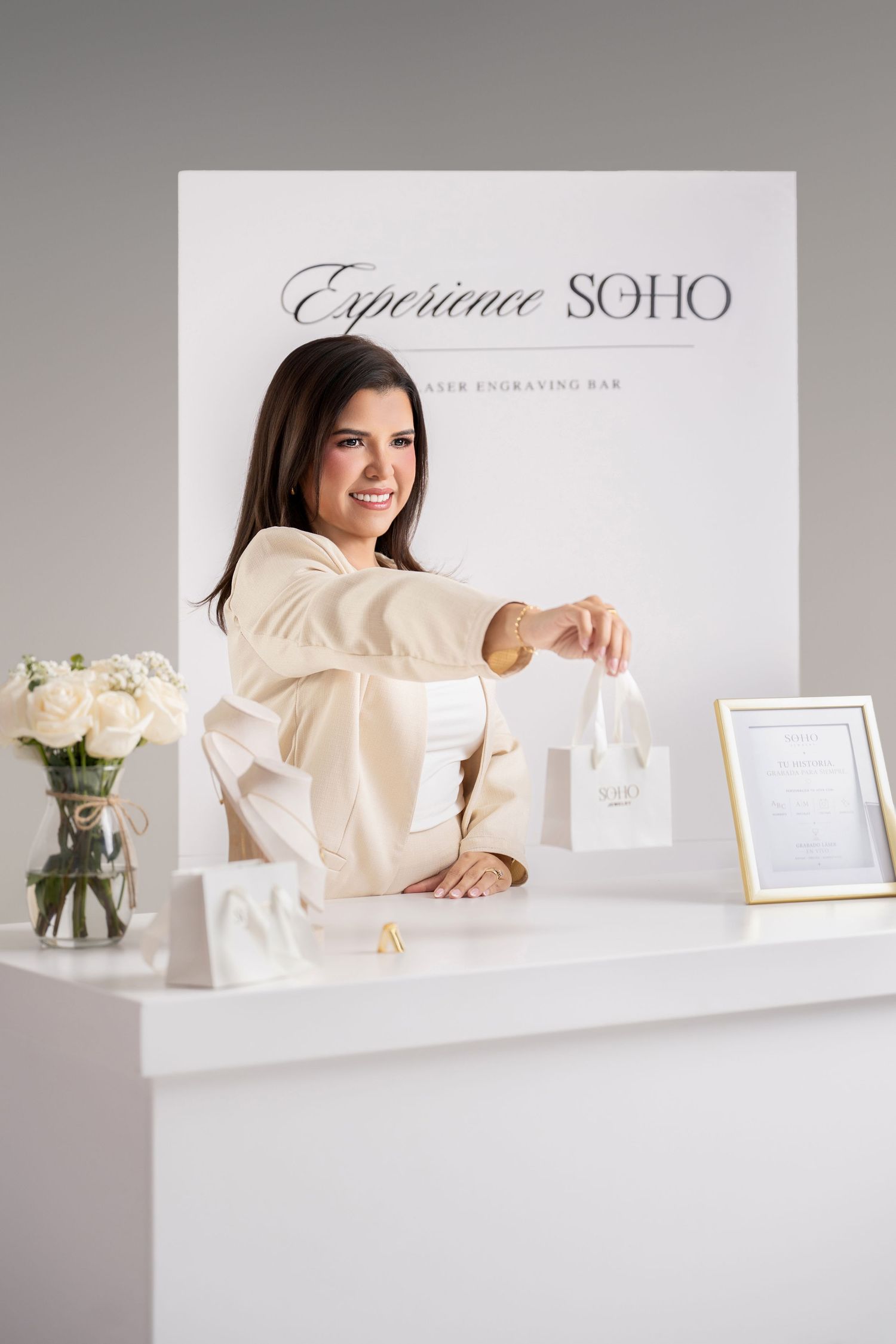 Smiling brunette woman at white retail counter handing a small shopping bag at Experience SOHO engraving bar.