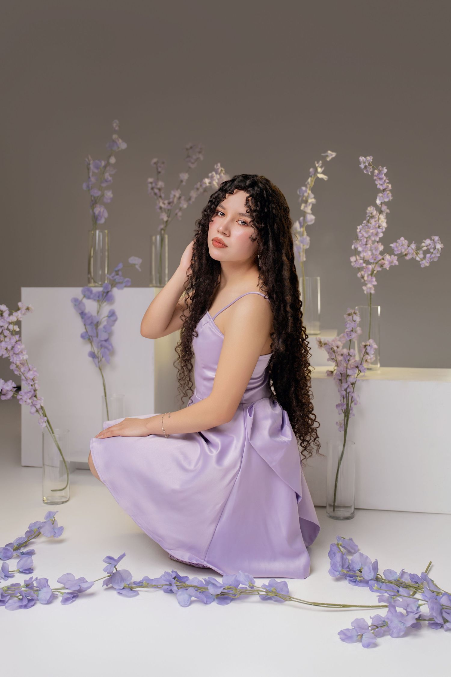 Young woman with long curly hair in a lavender satin dress sits among purple flowers and glass vases on a gray background.