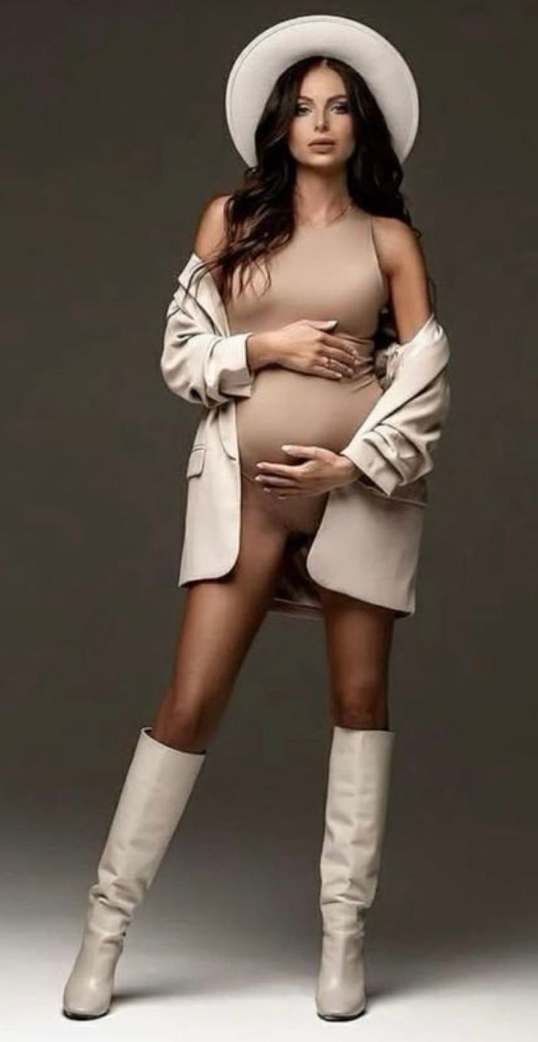 Pregnant woman in beige bodysuit, cream blazer, white hat and knee-high boots posing for a maternity photo shoot.