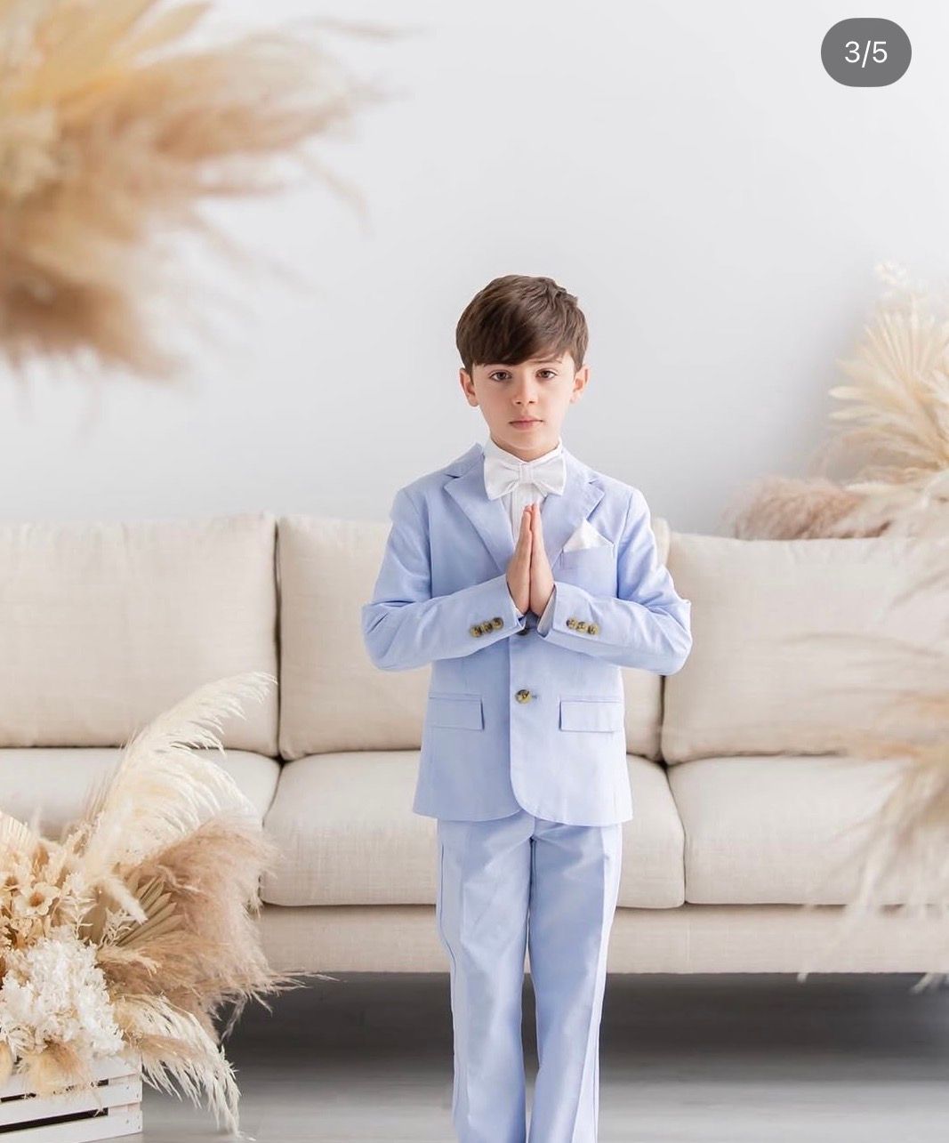 Boy in light blue suit with bow tie, hands in prayer pose, standing in front of beige sofa with pampas grass decor.