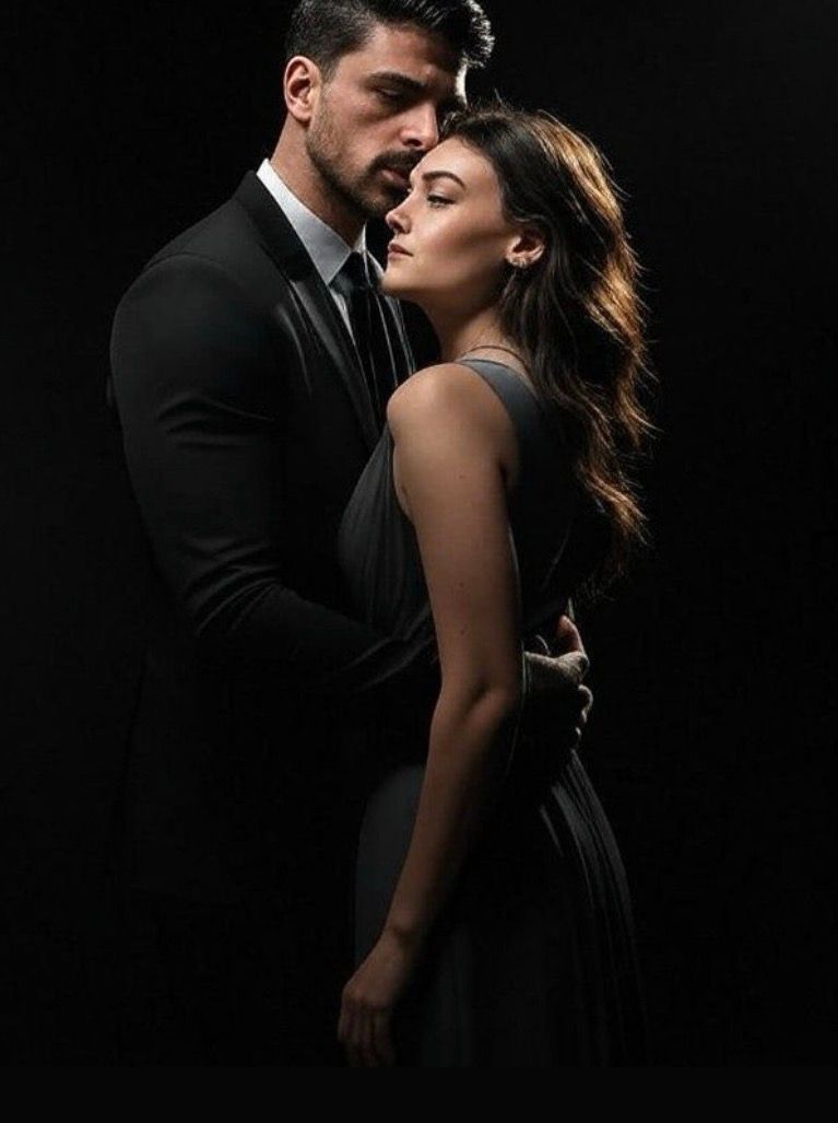 Elegant couple in black attire embrace intimately against a dark background in a romantic, dramatic portrait.