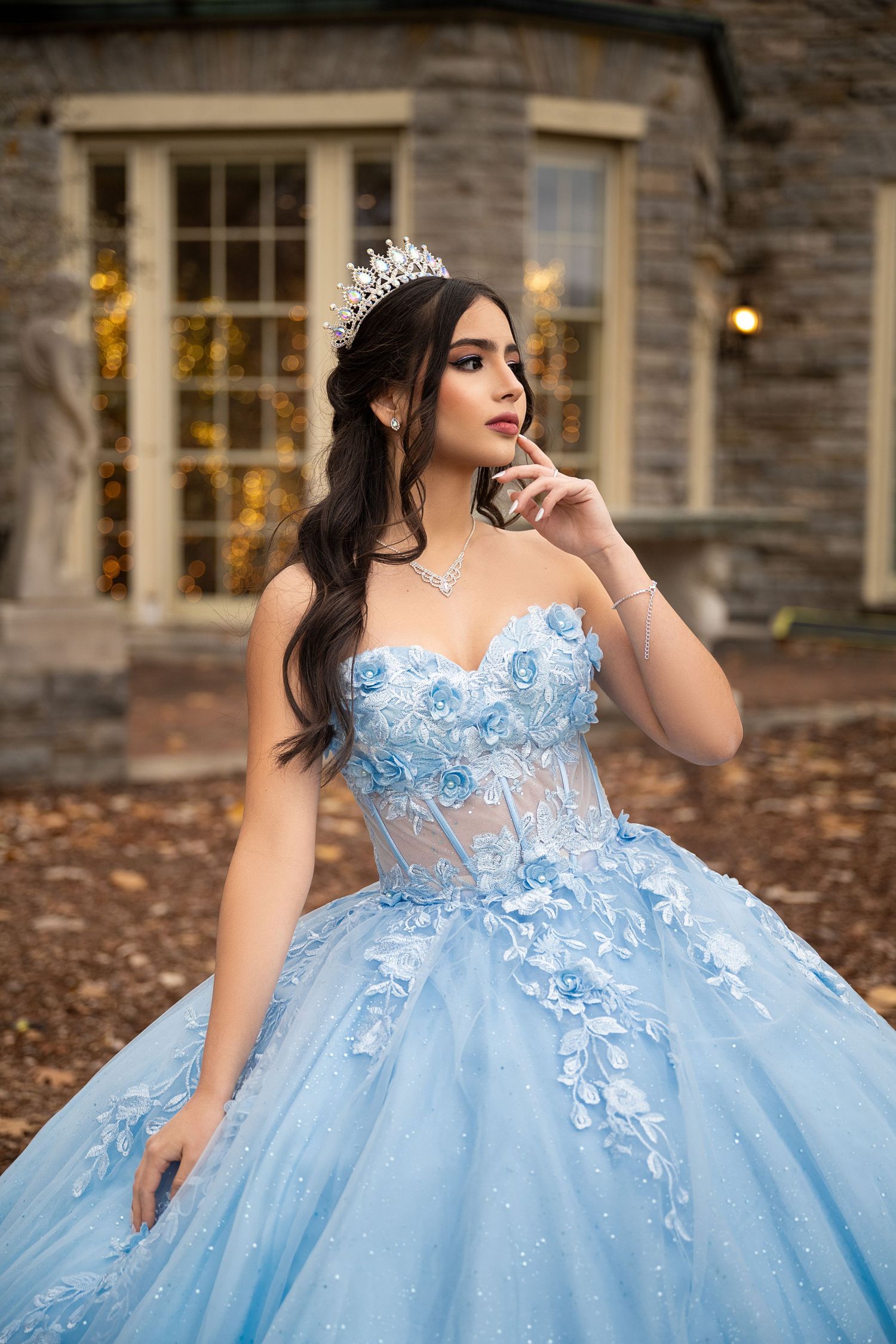 Elegant light blue ball gown with floral appliques and sheer corset bodice shown in autumn setting with stone building backdrop.