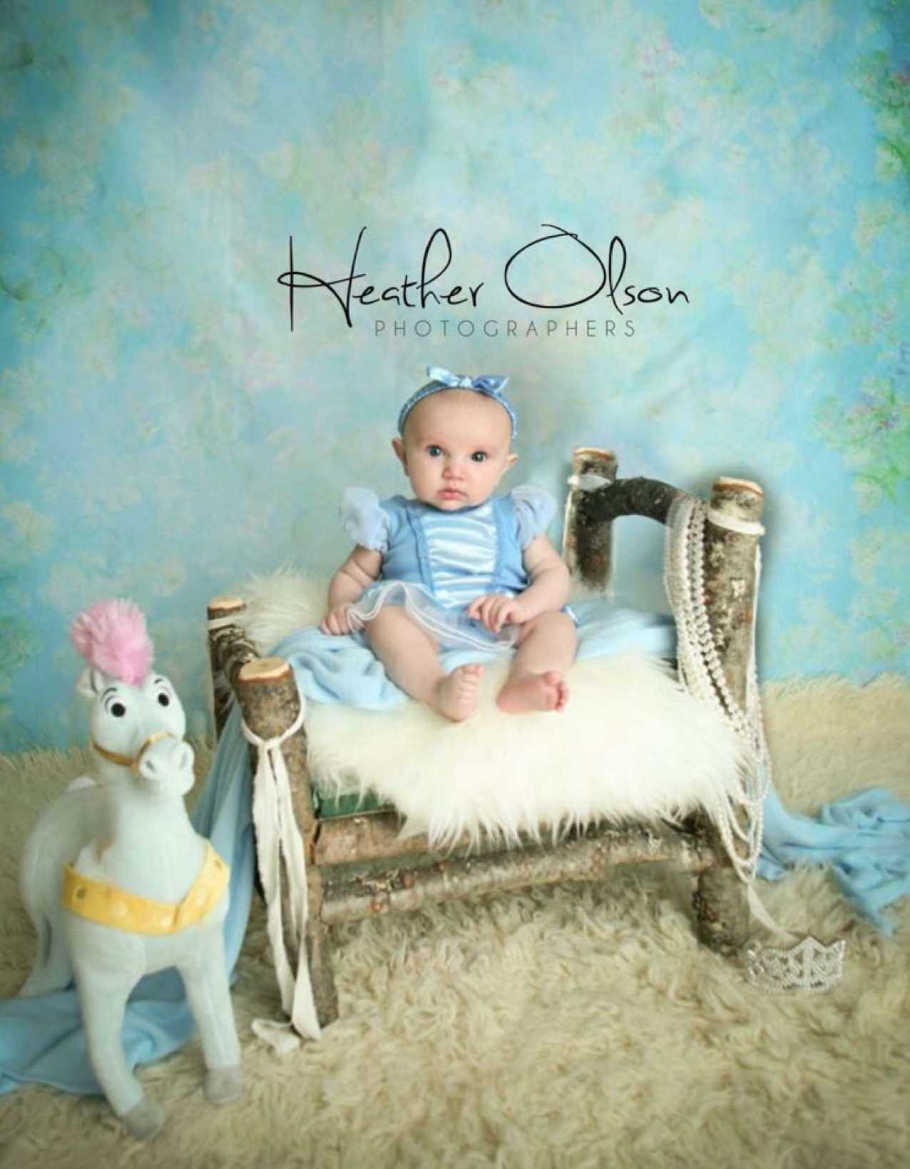 Baby in blue Cinderella outfit sits on fur-covered bench with toy horse, professional photo by Heather Olson.