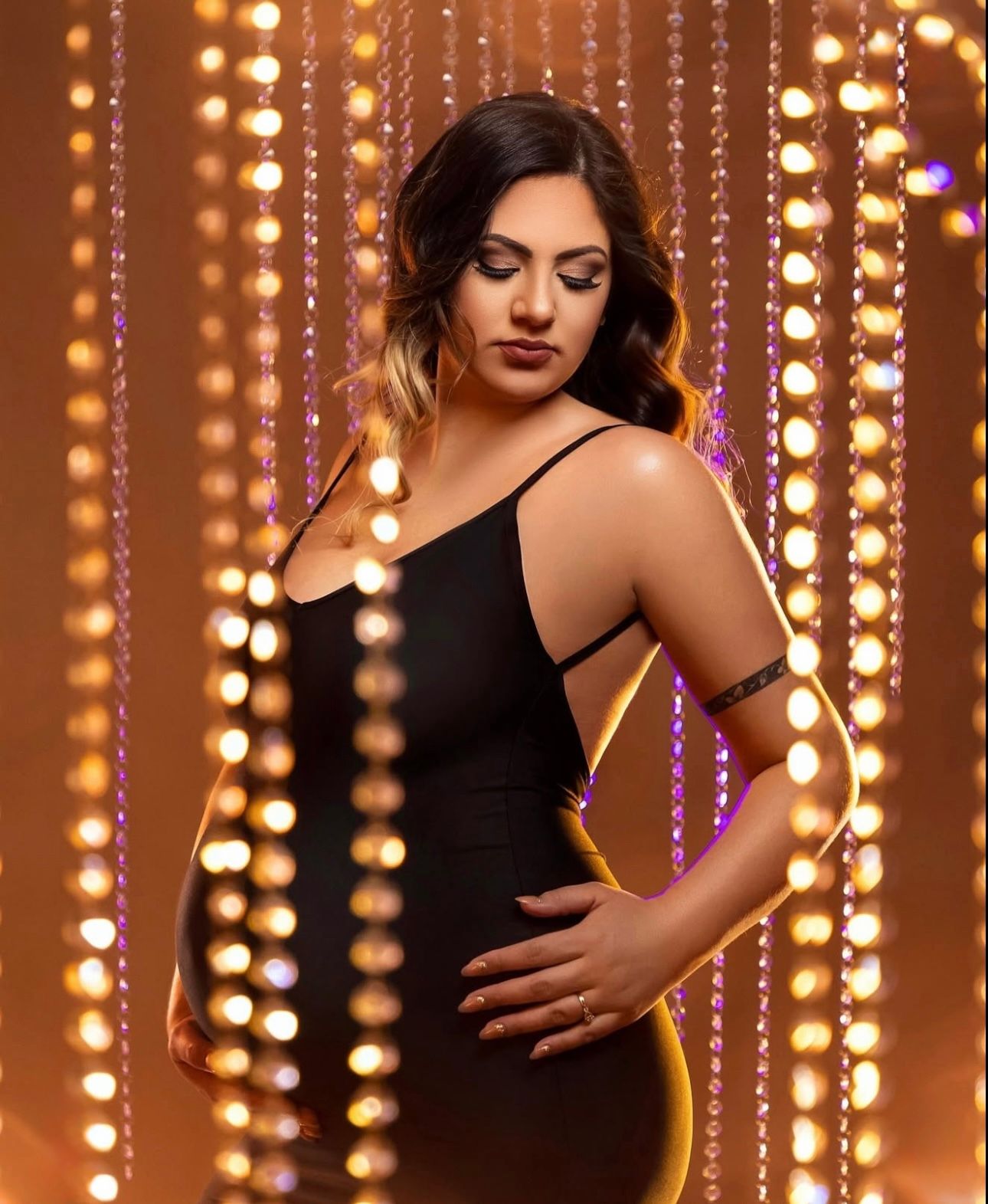 Woman in black spaghetti strap dress posing elegantly against a glowing bokeh light curtain backdrop.