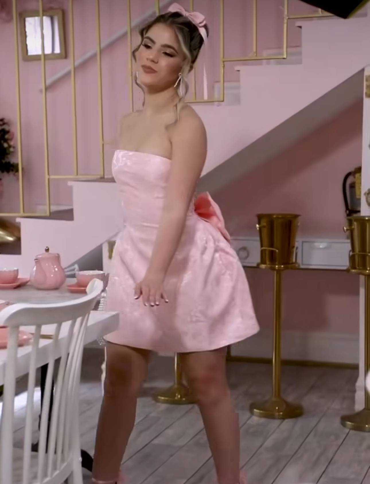 Young woman posing in a strapless pink mini dress with bow detail in a pink-themed room with gold accents.