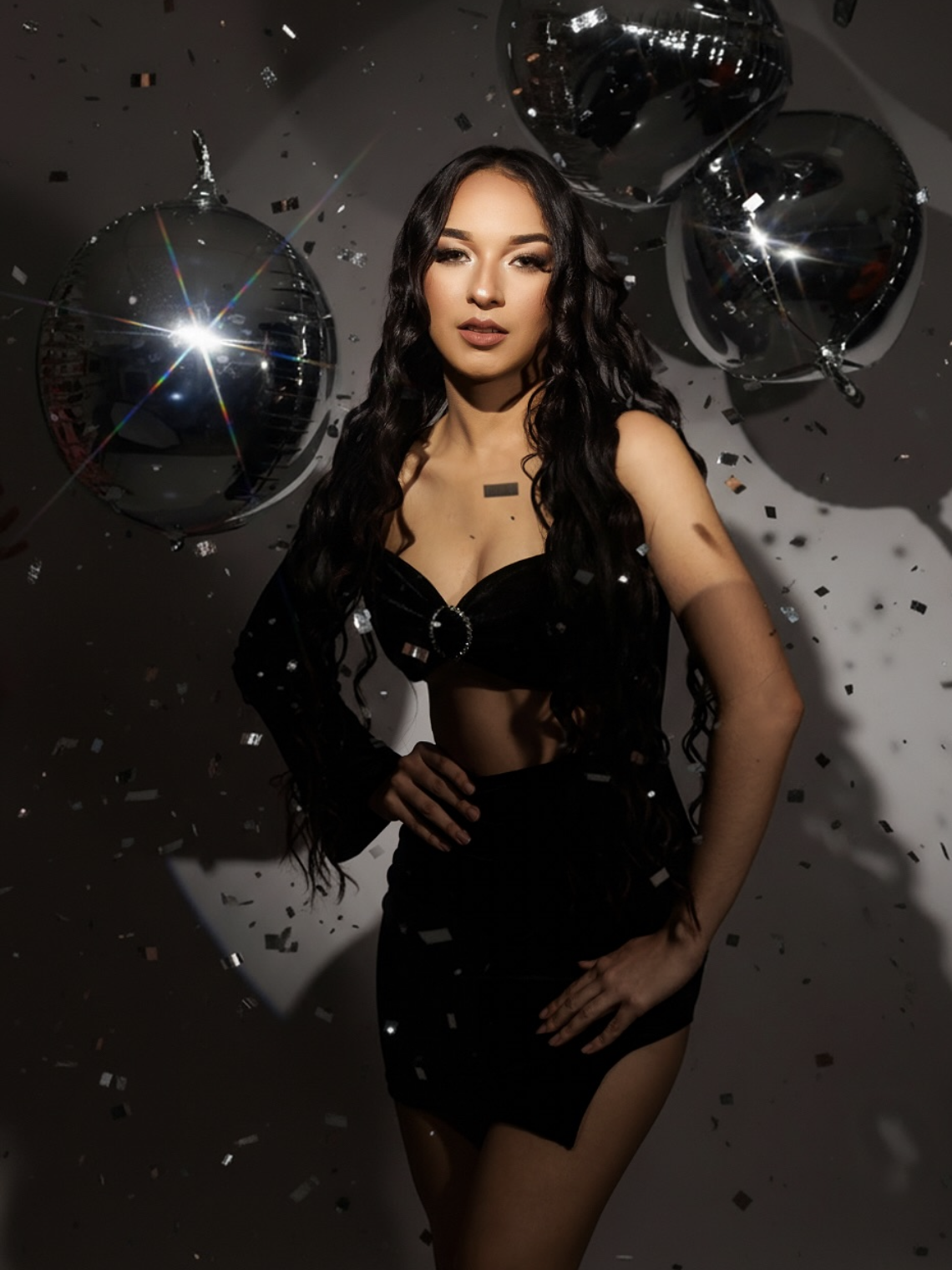 Woman in black outfit poses with disco balls and confetti in a glamorous party-themed photo shoot.