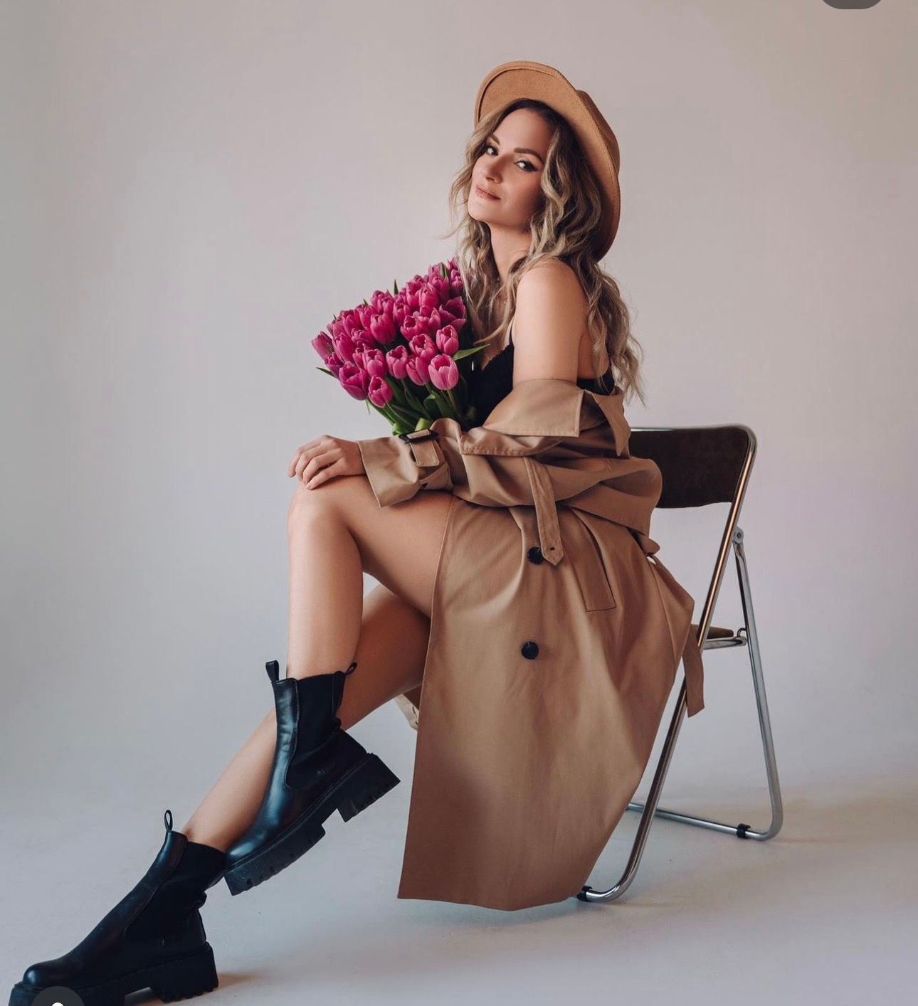 Woman in beige trench coat and beret sits on folding chair holding pink tulips, wearing black ankle boots.