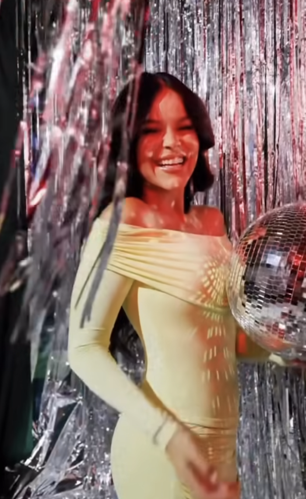 Smiling woman in yellow off-shoulder dress holding disco ball amid silver tinsel curtains at party.
