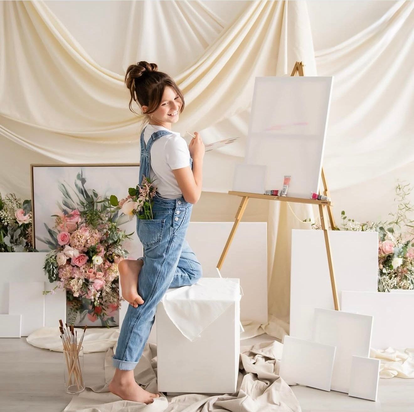 Young woman in denim overalls painting on canvas easel surrounded by white frames and pink floral arrangements in bright studio.