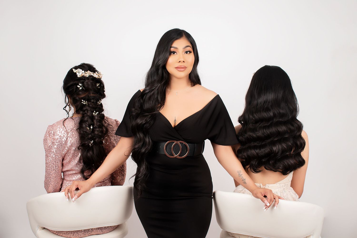 Three women in black dresses sitting on white chairs posing for glamorous photo.