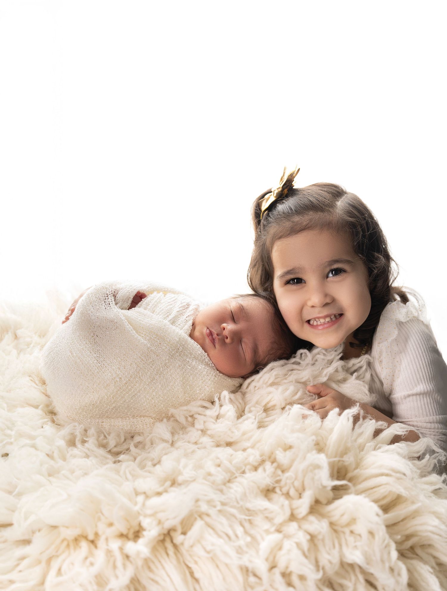 Sweet sibling moment on soft white fluffy rug with newborn baby wrapped in blanket.