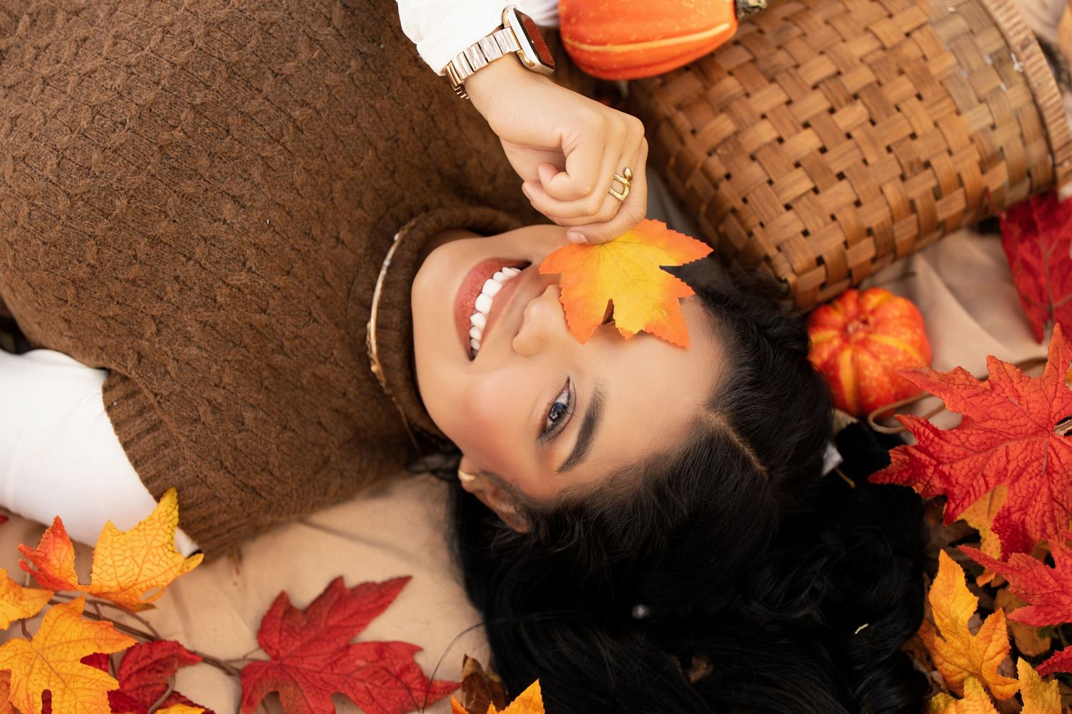 Woman smiling among autumn leaves, holding a maple leaf, with pumpkins and a wicker basket in a fall setting.