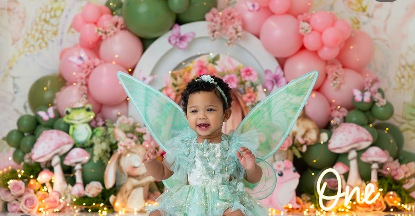 Baby girl in a mint fairy costume with wings celebrates her first birthday surrounded by pink and green balloon decorations.