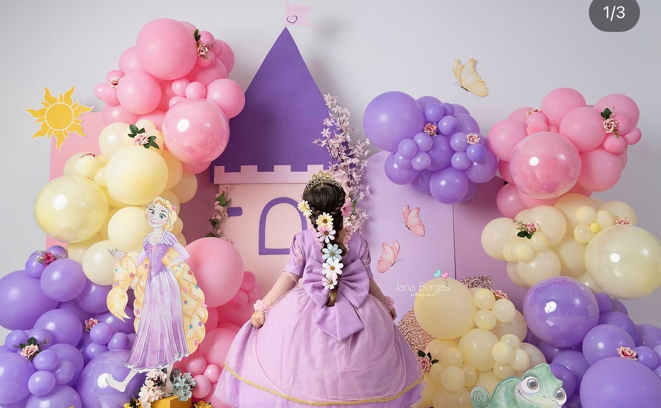 Girl in purple princess gown poses before a Rapunzel castle backdrop with colorful balloon garland decorations.