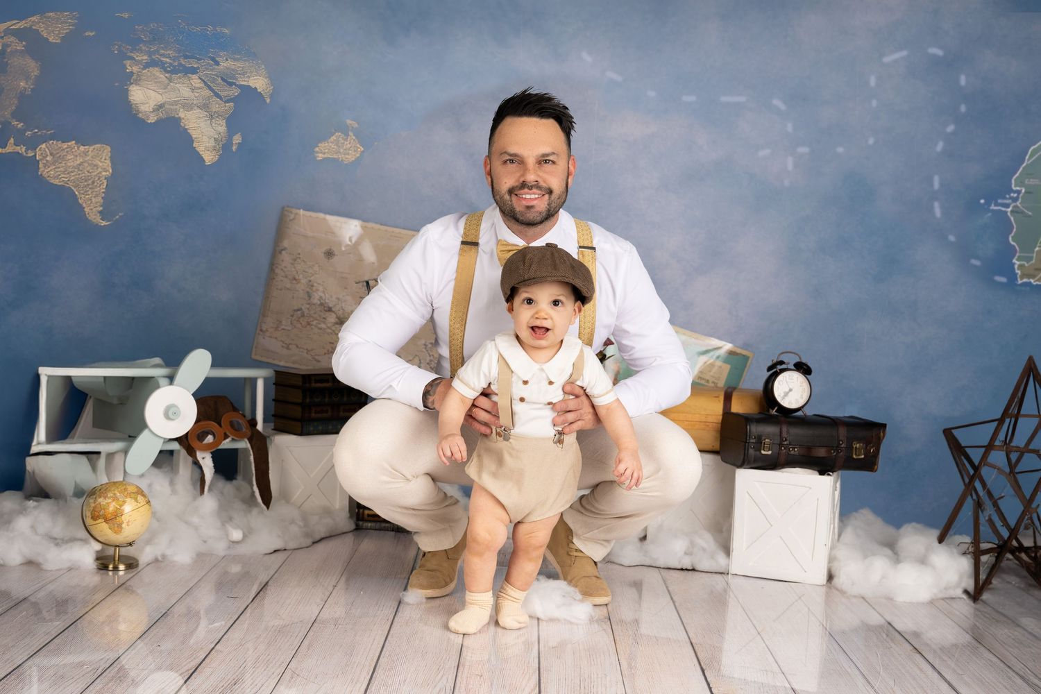 Father kneeling behind baby in suspenders and cap, posing in a travel-themed photo studio with map backdrop.