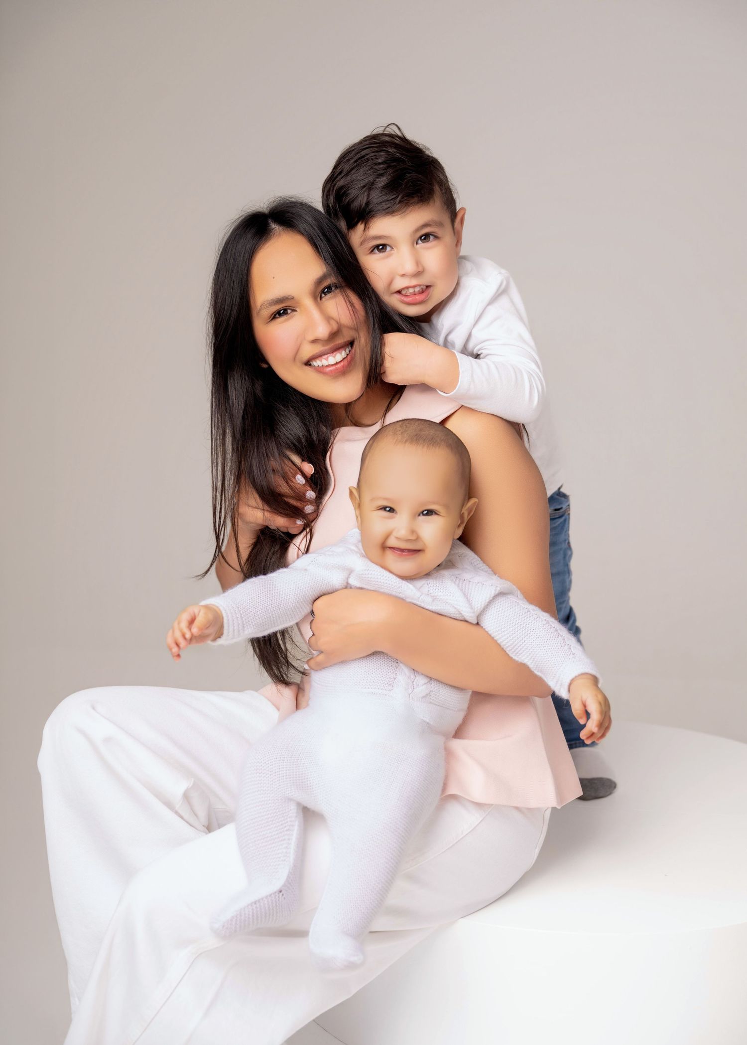 A smiling woman in white poses with two young children, a toddler boy and a baby, in a bright studio portrait.