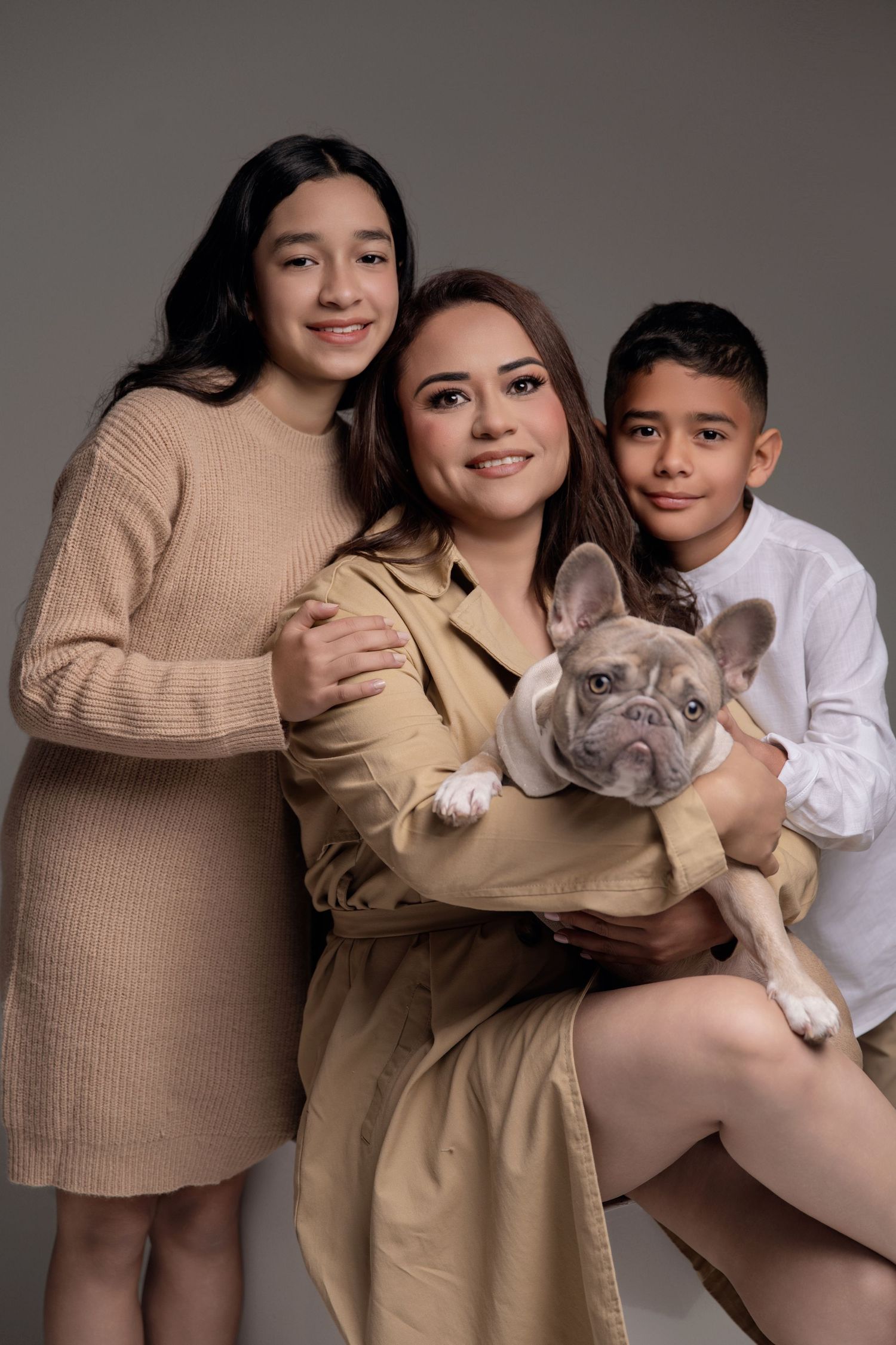 A woman holds a French Bulldog while posing with two children in neutral tones against a gray studio background.