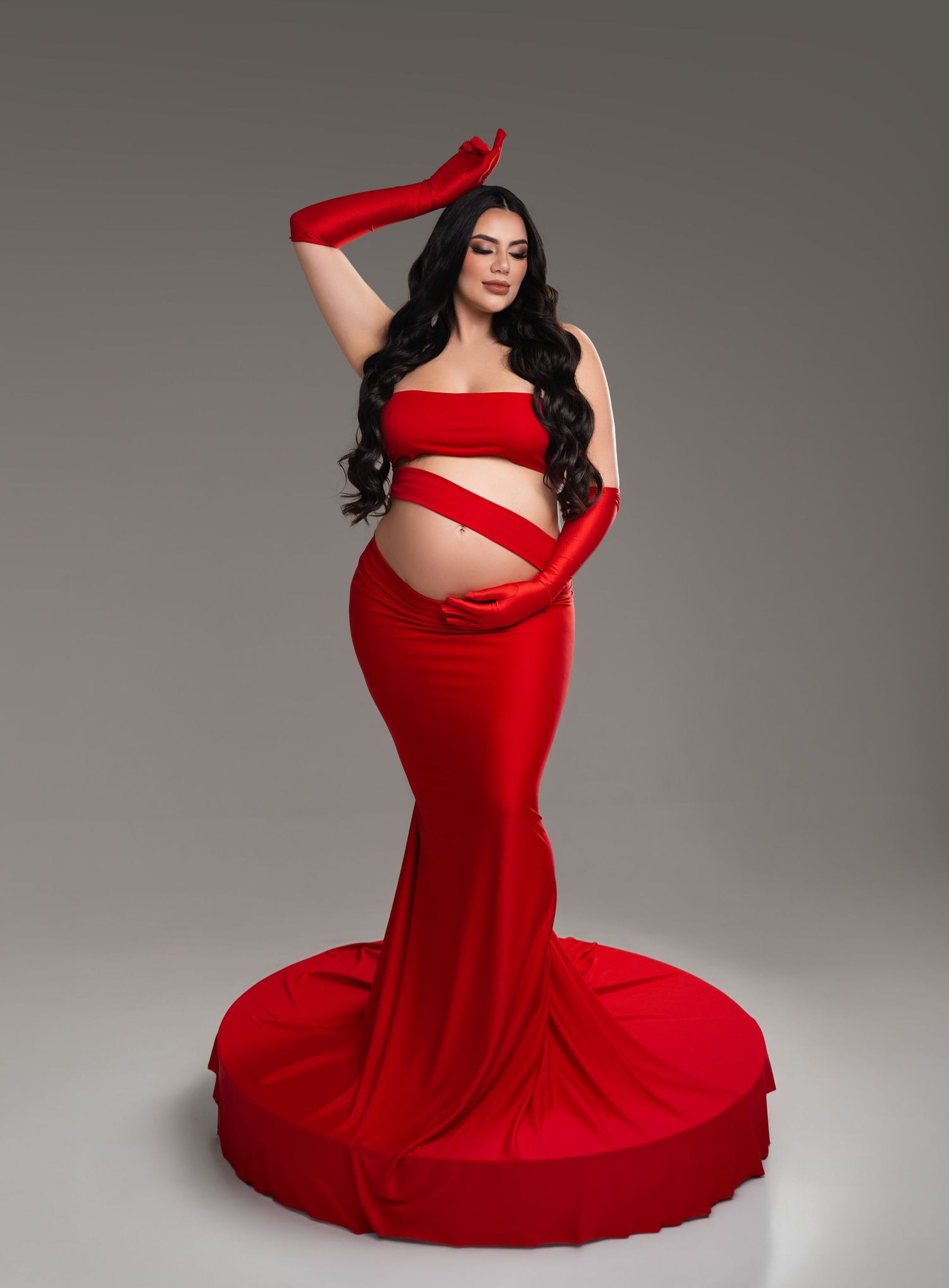 Pregnant woman in red strapless mermaid gown with matching gloves poses gracefully in a maternity photo shoot.