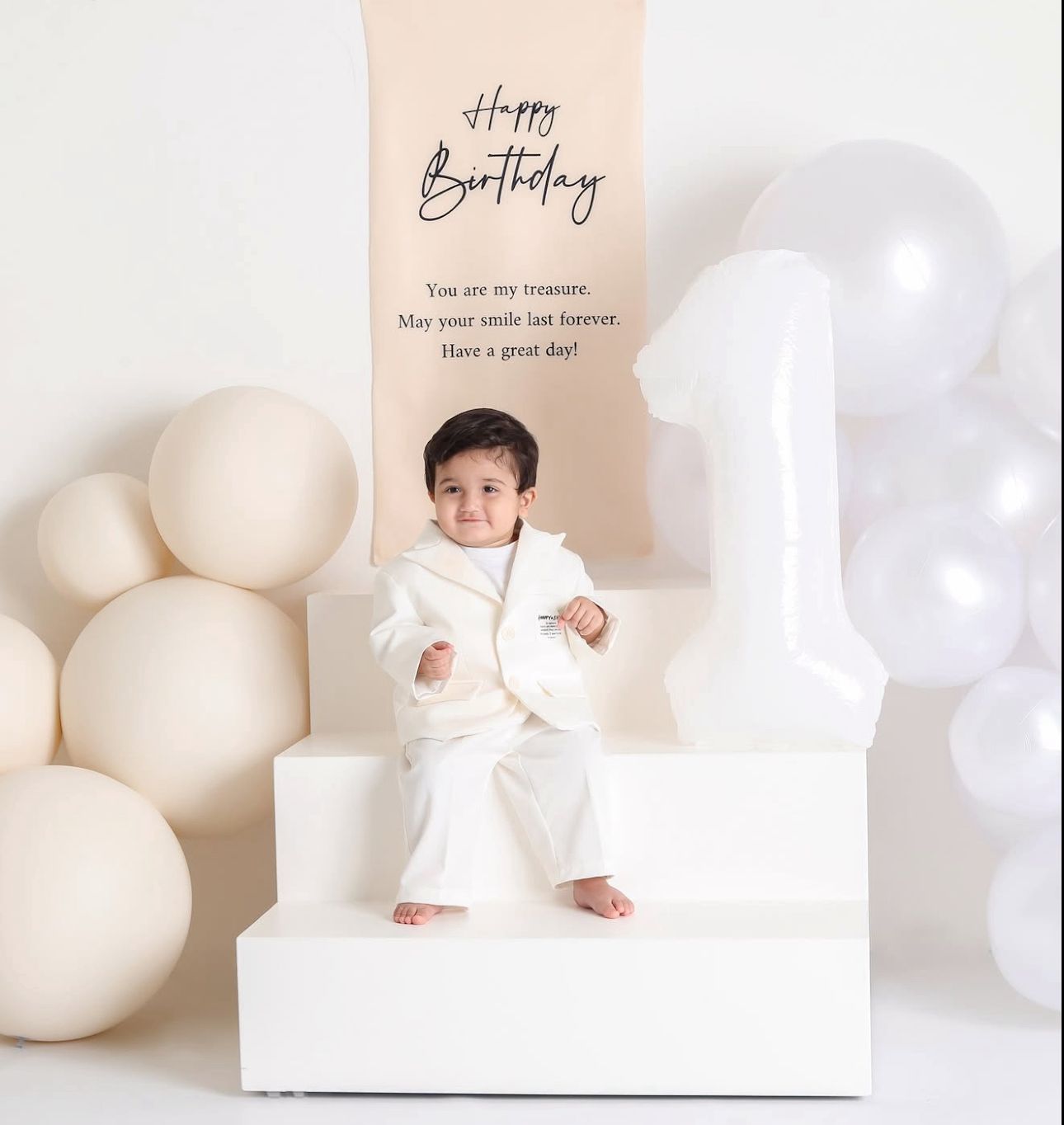 Minimalist birthday photo setup with white balloons and decorations against a light beige background.