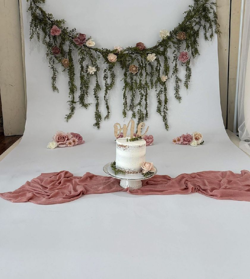 A romantic wedding cake display with draped greenery garland, pink fabric, and floral accents on a white backdrop.