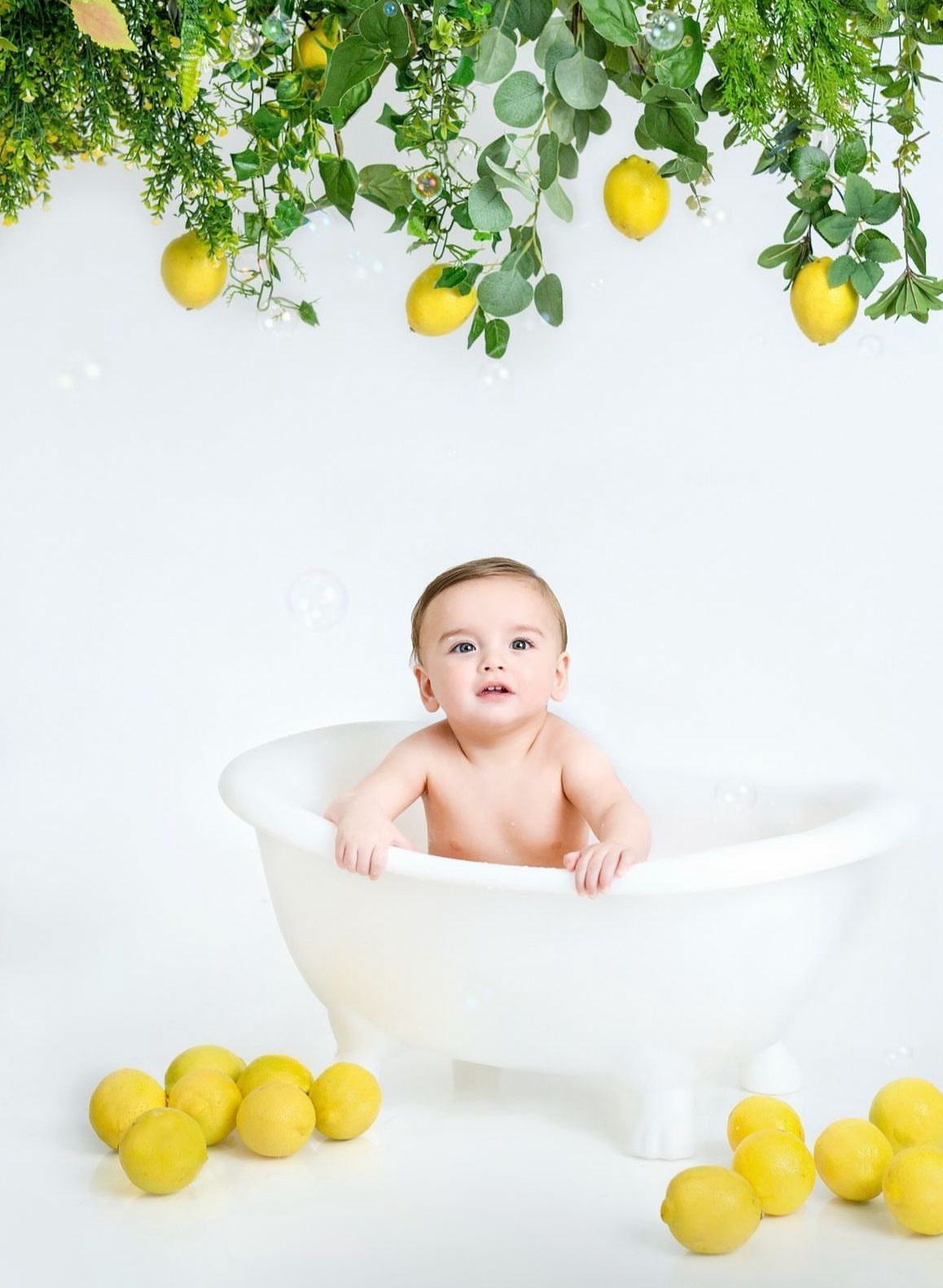 Fresh lemon-themed baby photography with white bathtub prop and hanging citrus foliage creating bright summer scene.