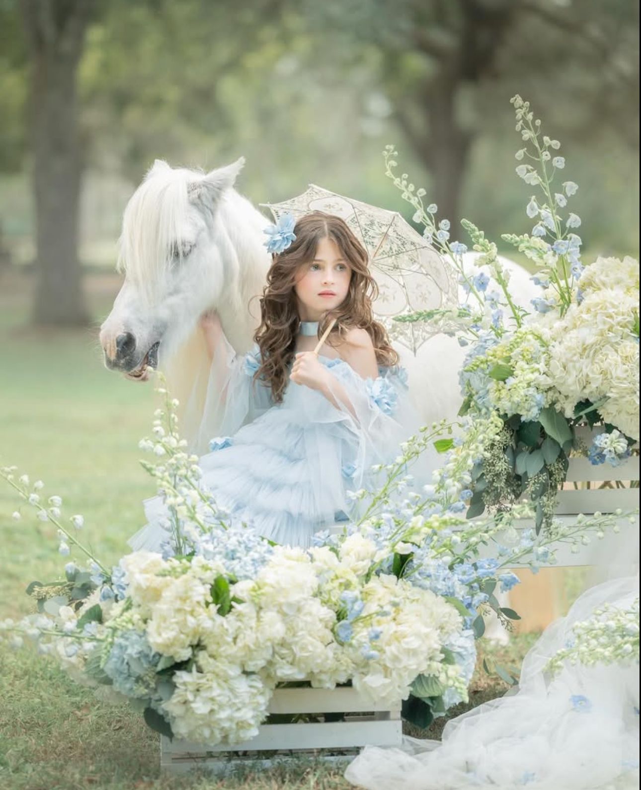 A young woman in a blue tulle dress holds a parasol beside a white horse surrounded by white and blue flowers outdoors.
