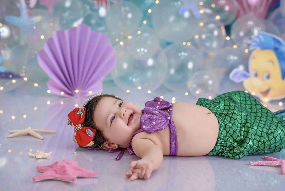 Baby girl in mermaid costume with purple top and green tail, smiling among seashells and starfish decorations.