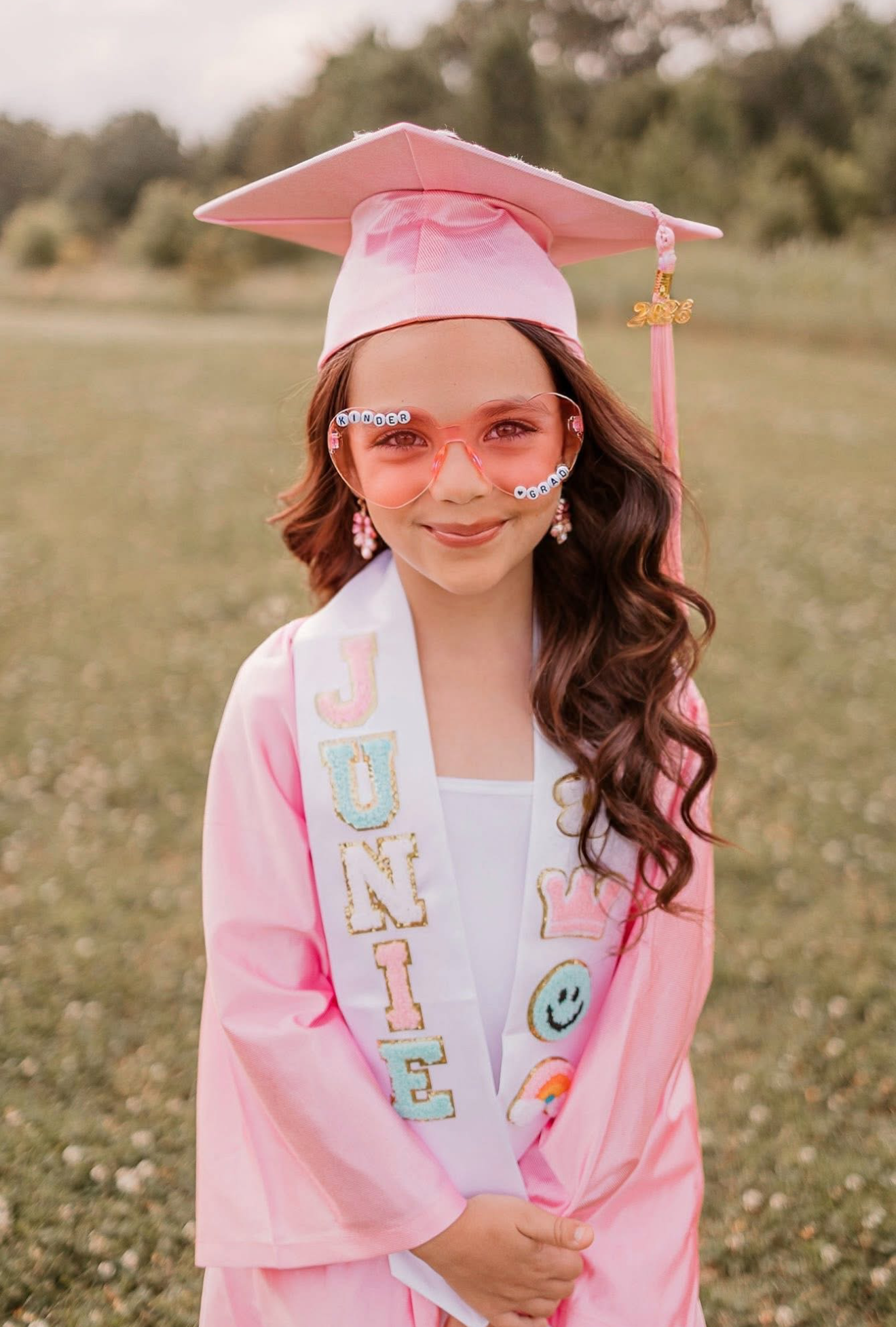 Young girl in pink graduation cap and gown with a personalized smiley face sash posing outdoors.