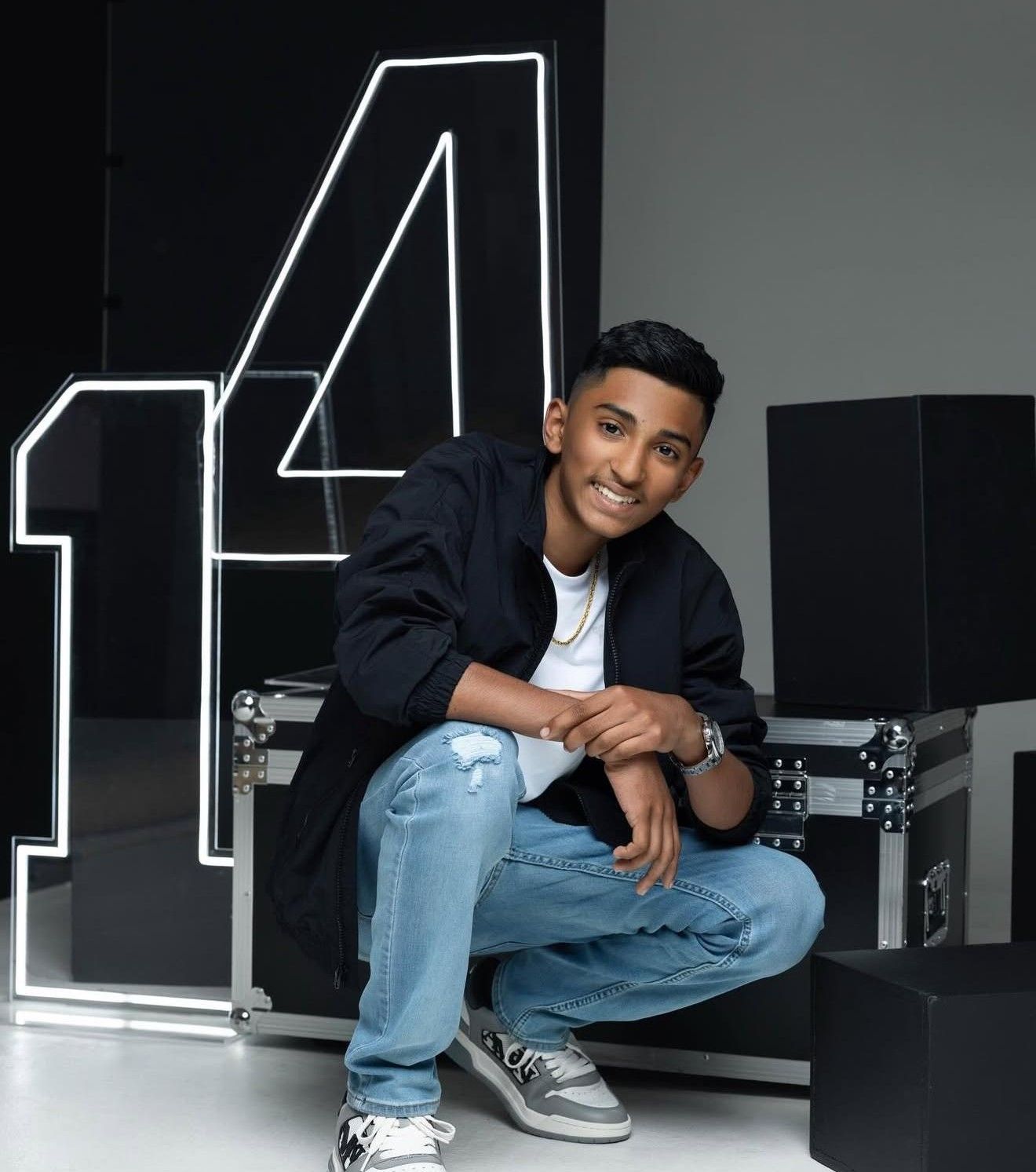Smiling young boy in blue jeans and black jacket crouching in front of large number 14 display.
